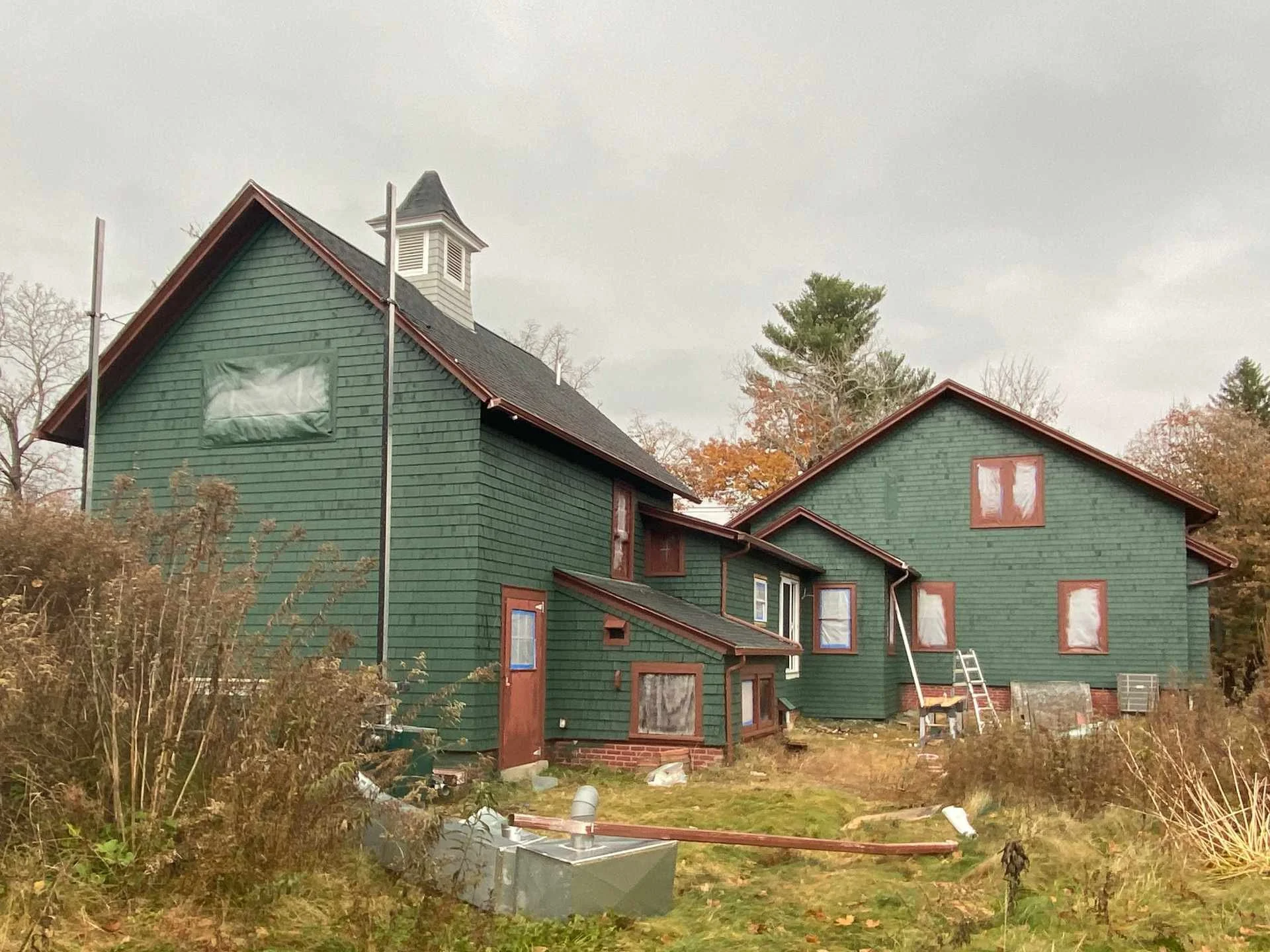 A green house with a steep dark roof and multiple windows, some covered with plastic. The house has a small tower-like structure on top with a vent. The yard is cluttered with construction materials, tools, and debris. There are trees in the backgrou