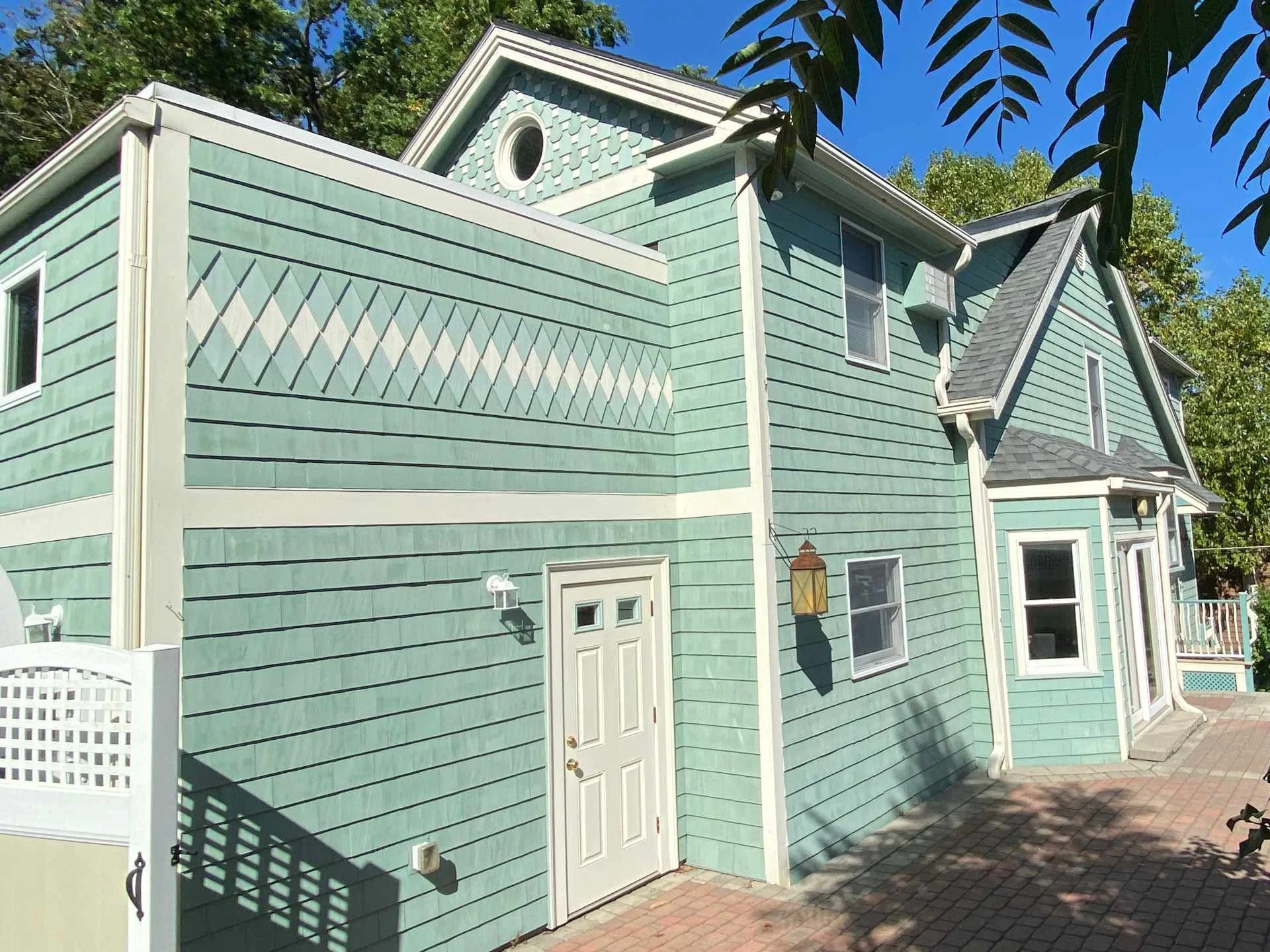A teal-colored house with multiple windows, a white door, and decorative shingles near the gable, set against a clear blue sky with leafy trees in the background.