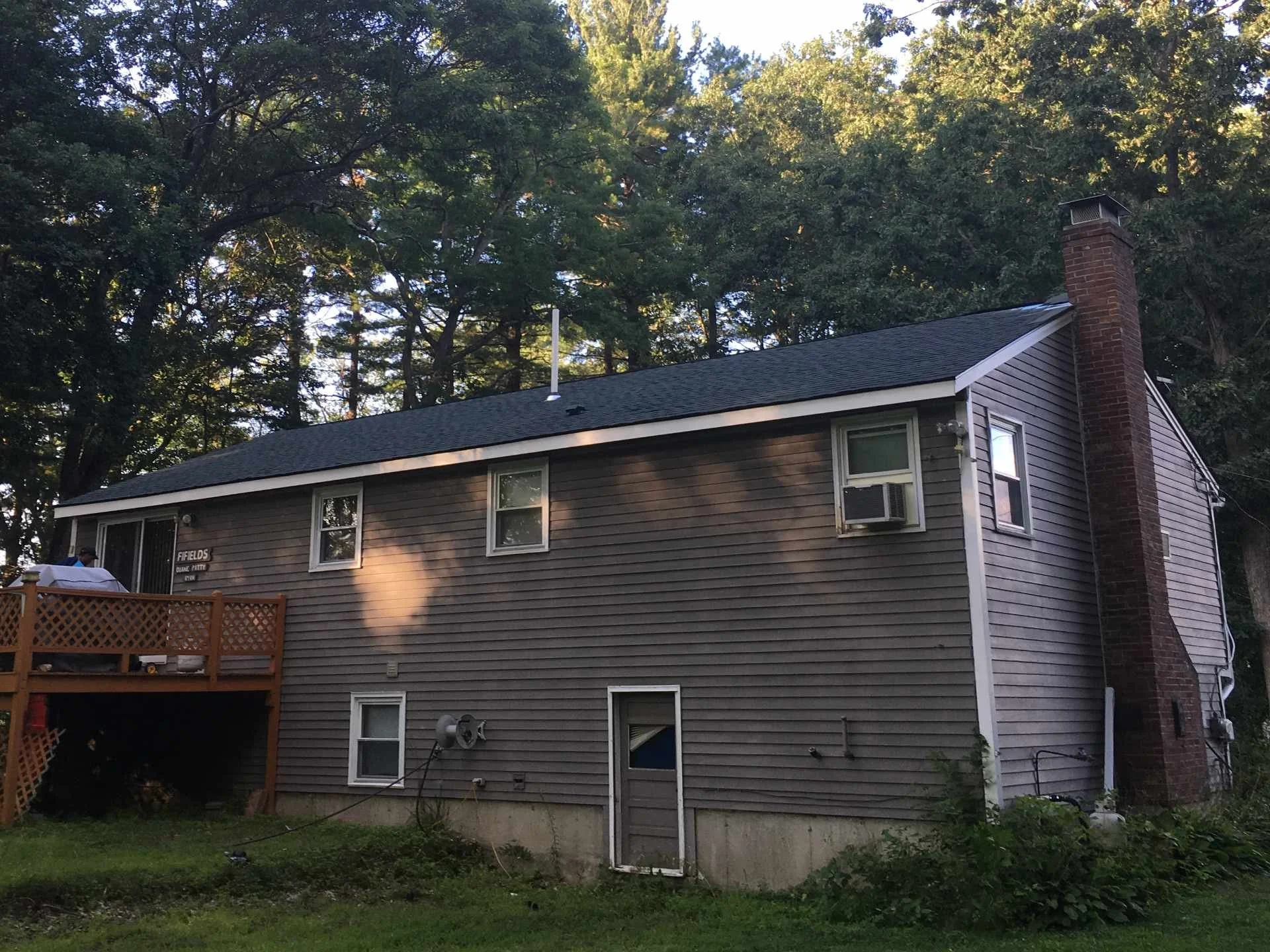 The photo depicts a newly finished house exterior renovation with gray siding, a chimney on the right side, a roof with black shingles, and multiple windows. There is a backyard with a wooden deck, some outdoor equipment, and a grassy area. Tall tree