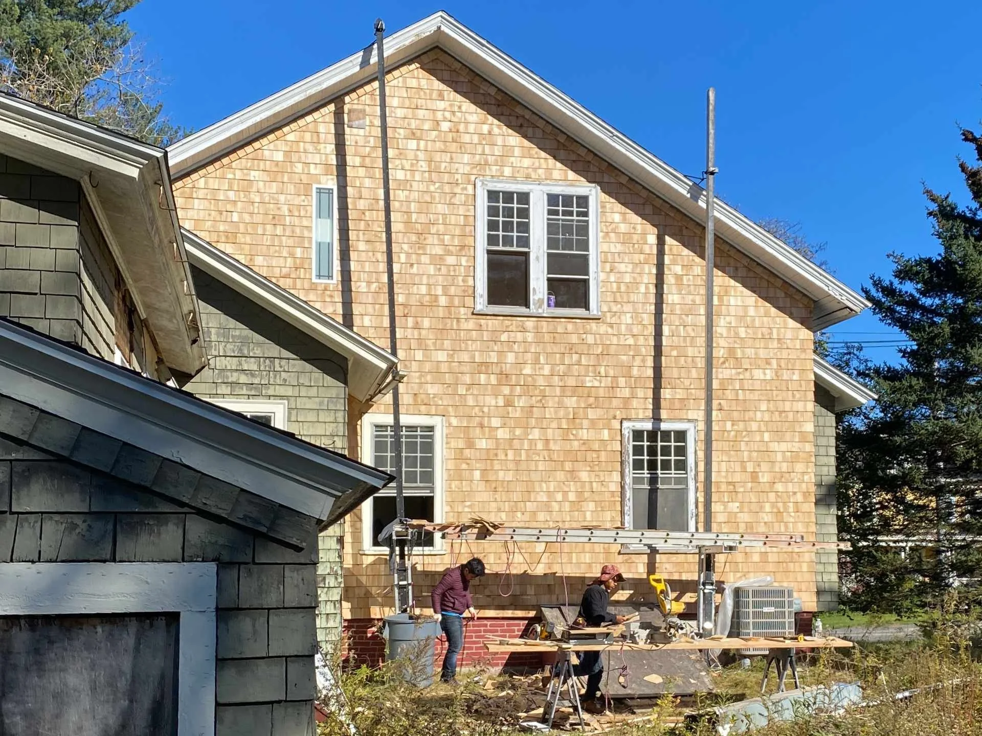 Two workers are building a house at a construction site with a wooden exterior and scaffolding. The house has multiple windows and is situated under a clear blue sky, with trees in the background.