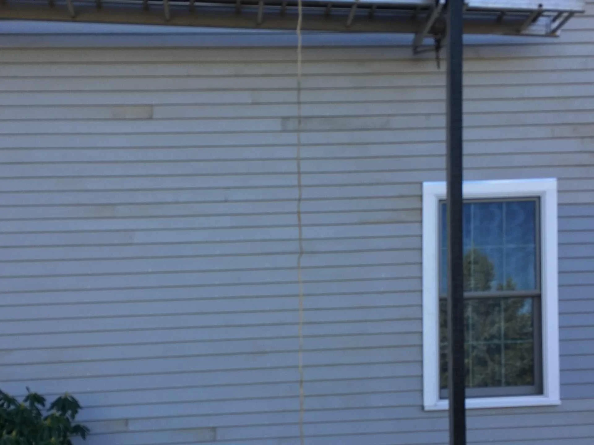 Side view of a house's exterior wall with beige horizontal siding, a window with white trim, and part of a black pole or post in front.