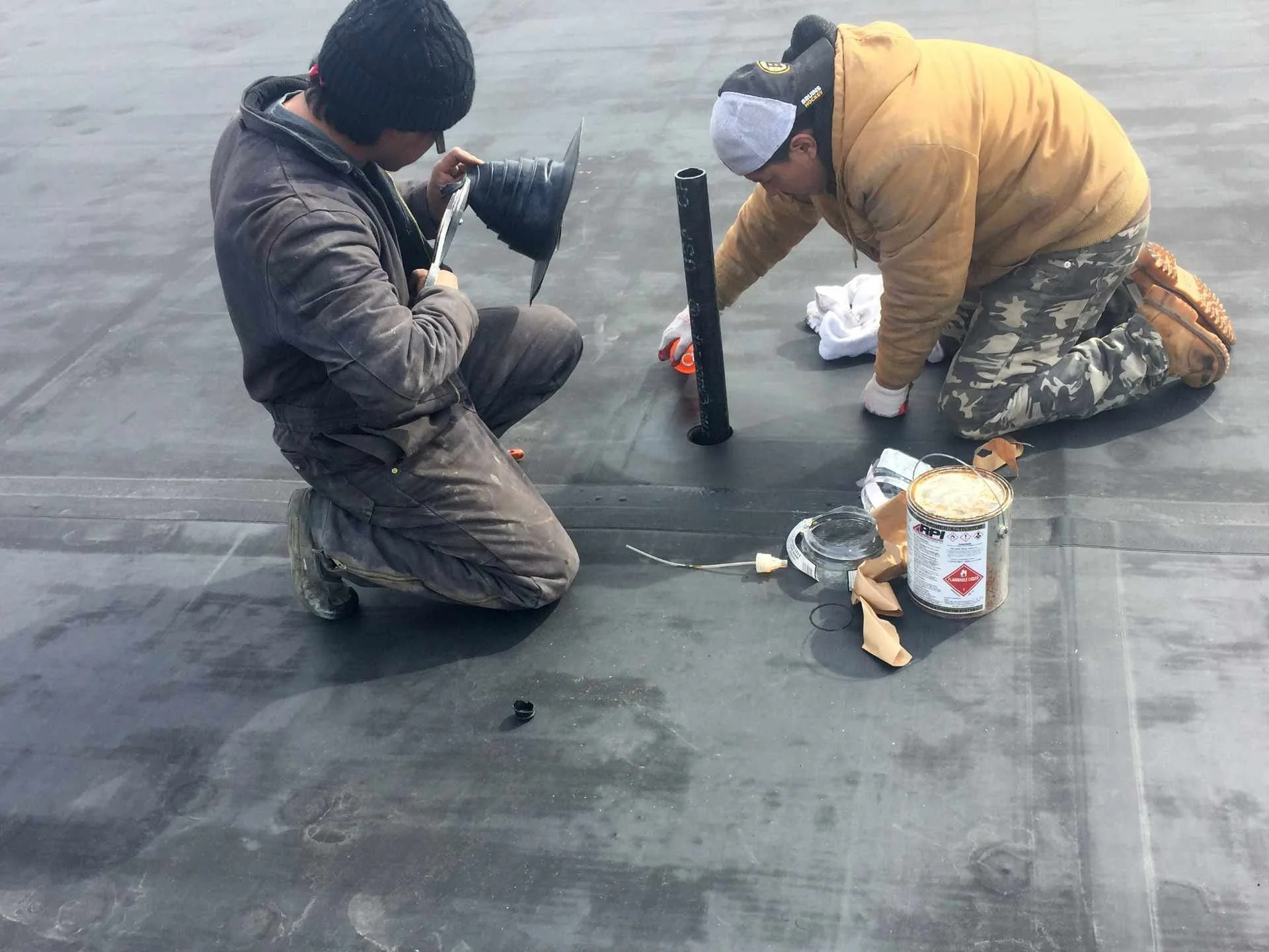 Two workers kneeling on a flat roof installing or repairing a waterproof membrane. One worker holds a trowel and a rubber boot, while the other uses a pipe for plumbing or venting. Various tools, a can of waterproofing material, and towels are scatte