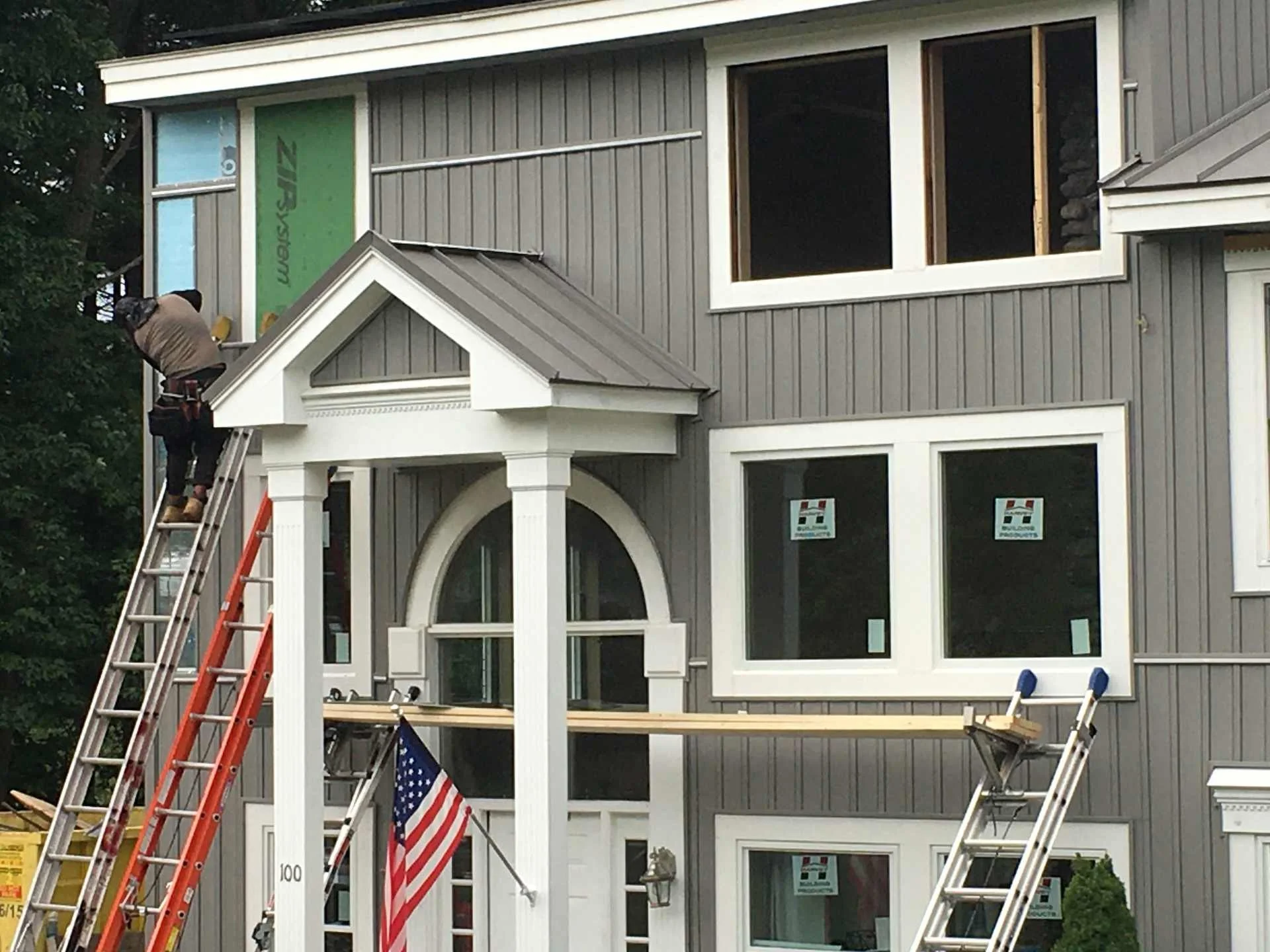 A construction worker is standing on a ladder working on the upper part of a gray house with white trim. The house has large windows and a covered front porch with white columns. An American flag is attached near the porch, and other ladders are visi