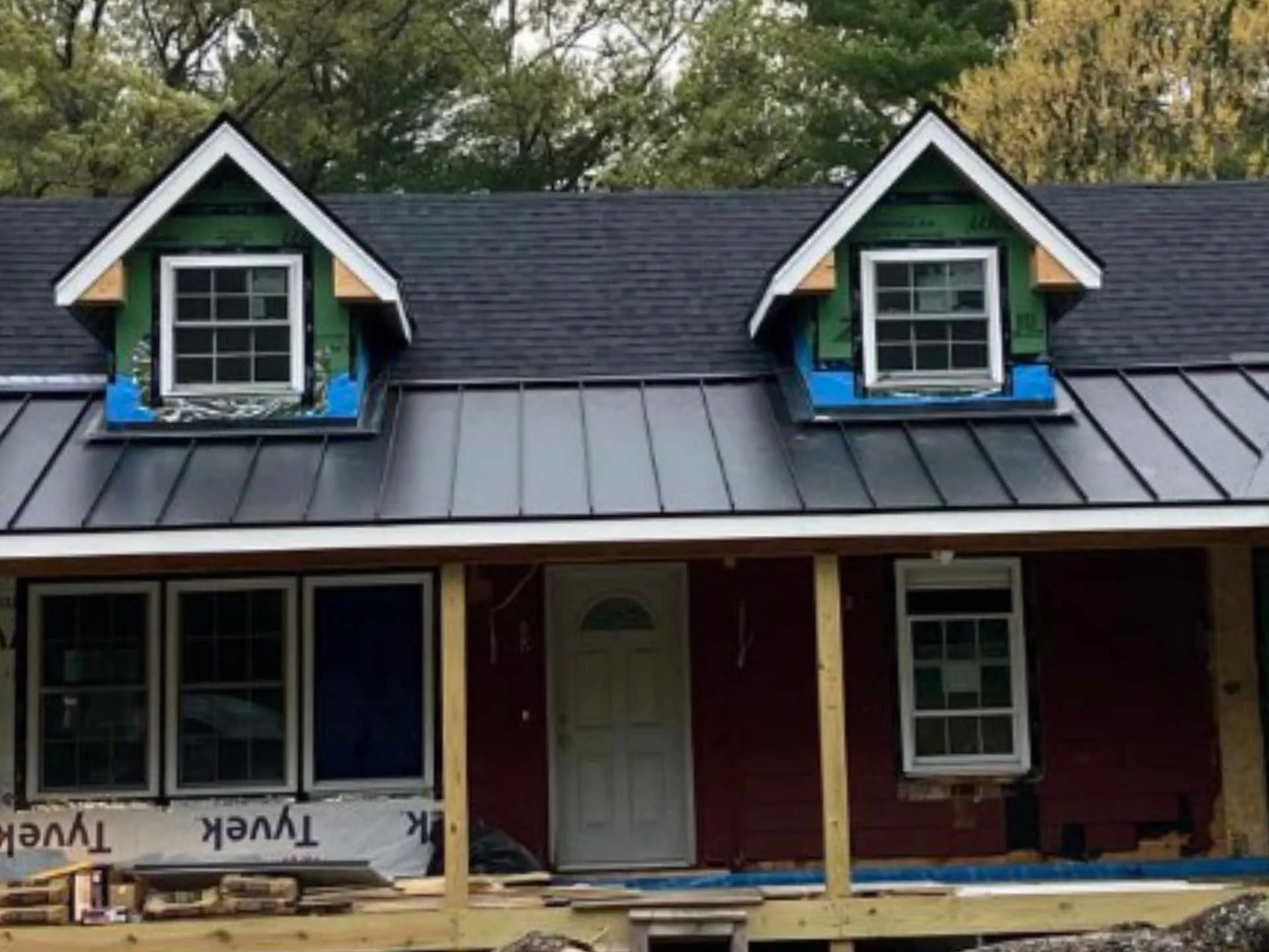 Under construction house with dormer windows, black asphalt roof shingles, black standing-seam metal panels, and an unfinished front porch in Chelmsford, MA. Trees are visible in the background.