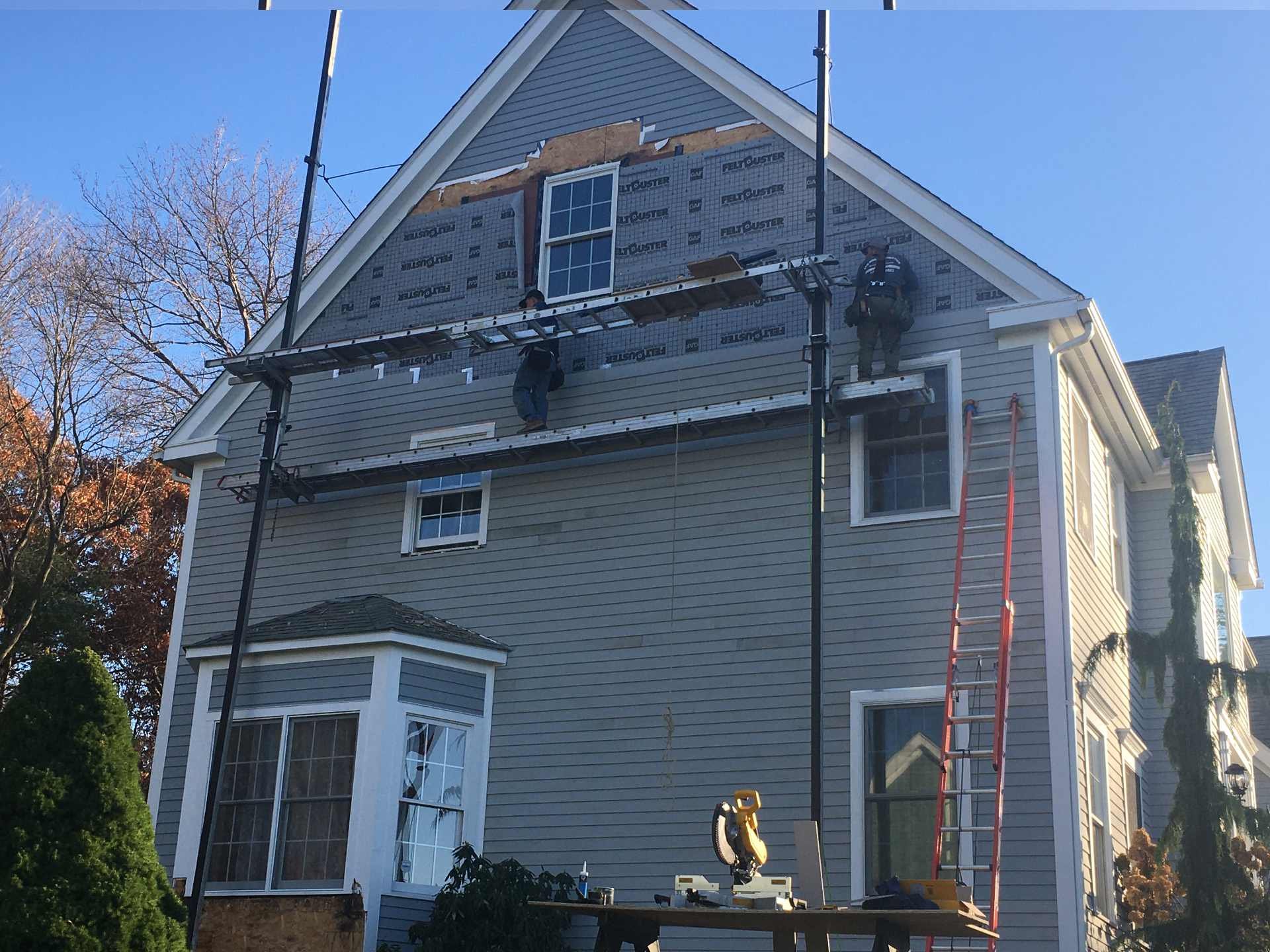 Construction workers installing siding on the upper exterior of a multistory house. Scaffolding is set up for their work, and tools are visible on a workbench in front of the house.
