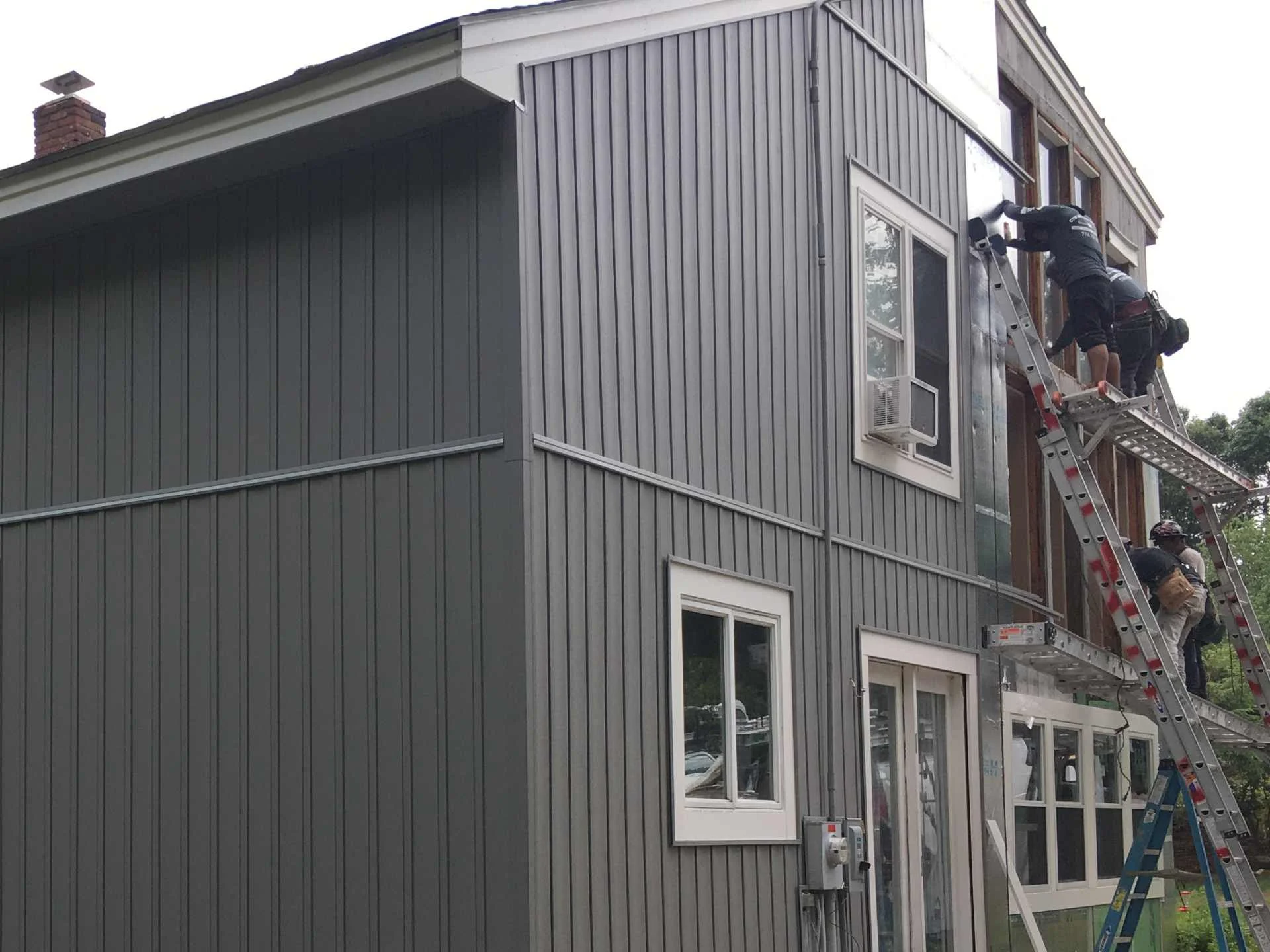Workers on a ladder installing vinyl board and batten siding after installing large windows on a multi-story house in Fitchburg, MA.