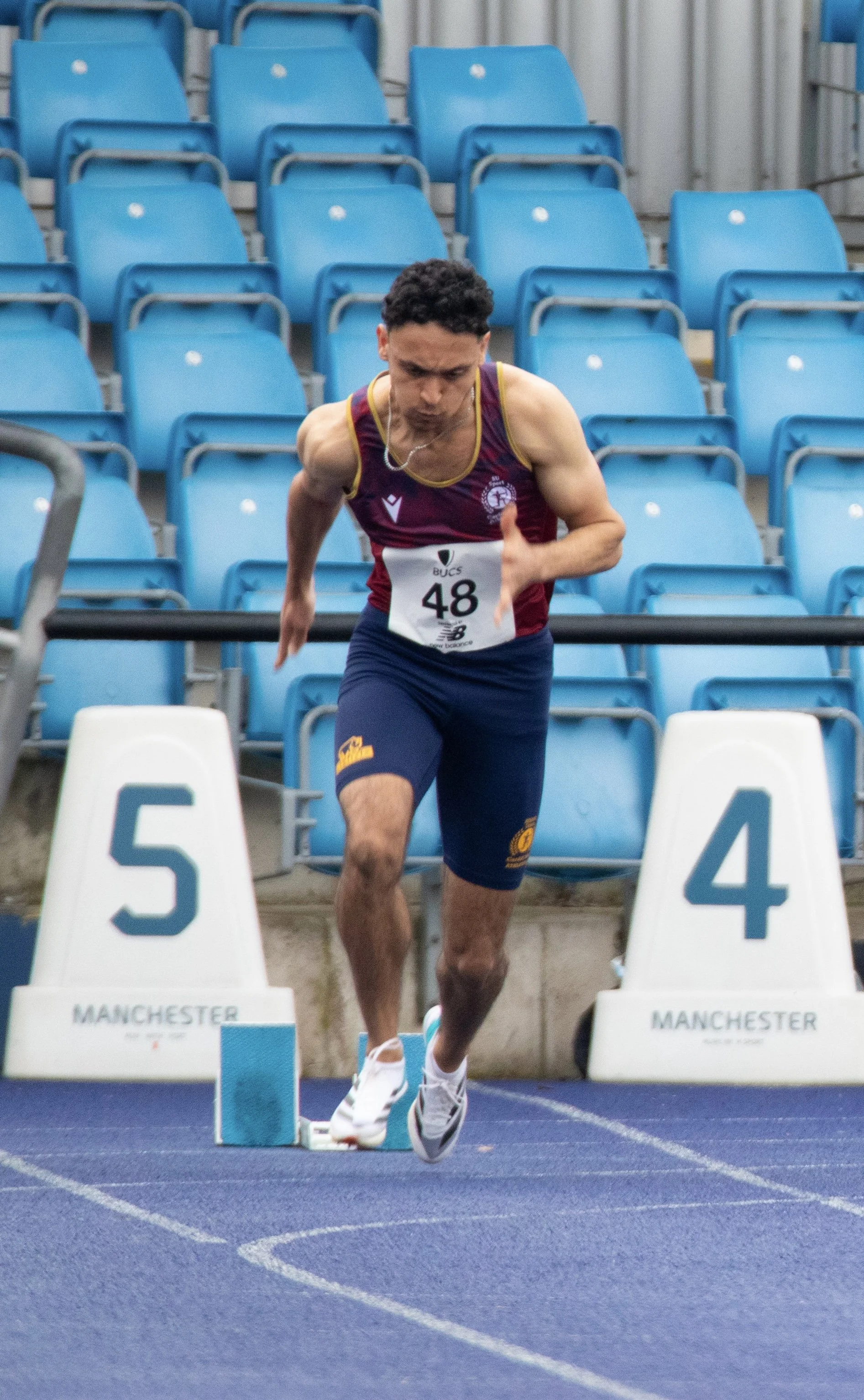 A male athlete running on a blue track during a race, with blue stadium seats in the background. He wears a maroon tank top, navy shorts, and white running shoes, with a bib number 48. Starting blocks labeled 'MANCHESTER' are in front of him.