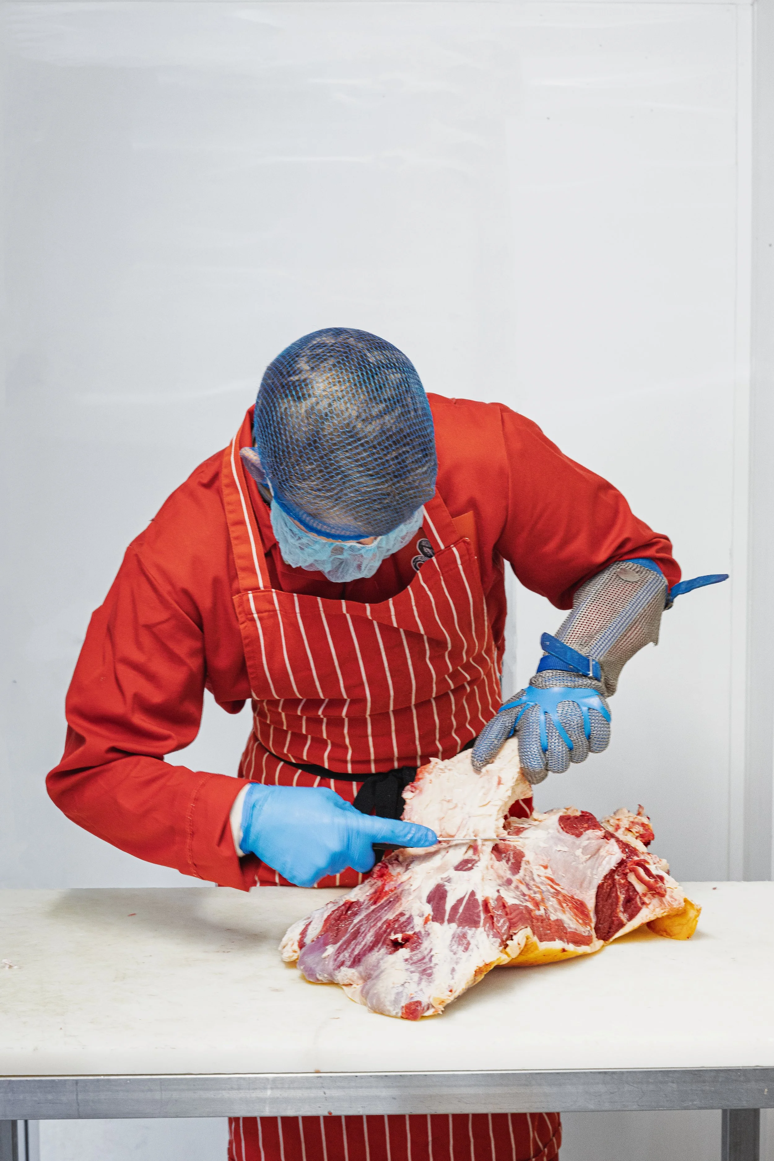 A person wearing a red apron, blue gloves, and a face mask is cutting raw meat on a white work surface. The background is plain white.