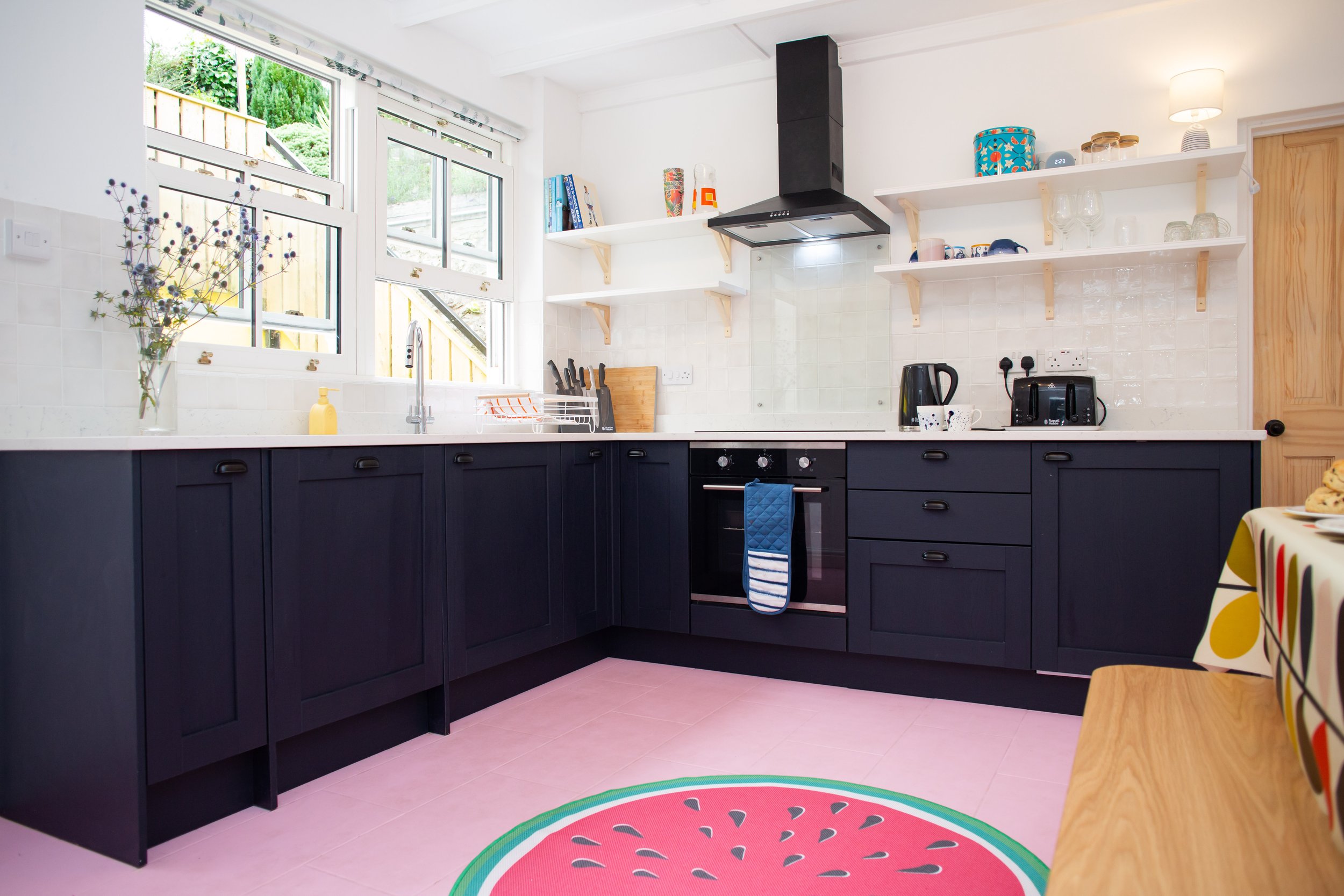 Kitchen with black cabinets, white countertop, gold faucet, oven, coffee maker, potted plants, butterfly artwork, and wall shelves with books and decorative items.
