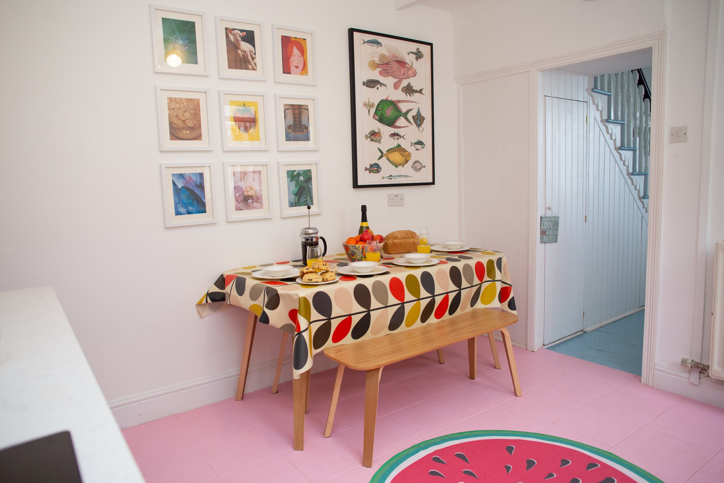 A cozy dining area with a small round table set for two, featuring two green chairs with sunflower-patterned cushions, a white table lamp, two glasses of rosé wine, and a backdrop mirror reflecting part of the kitchen.