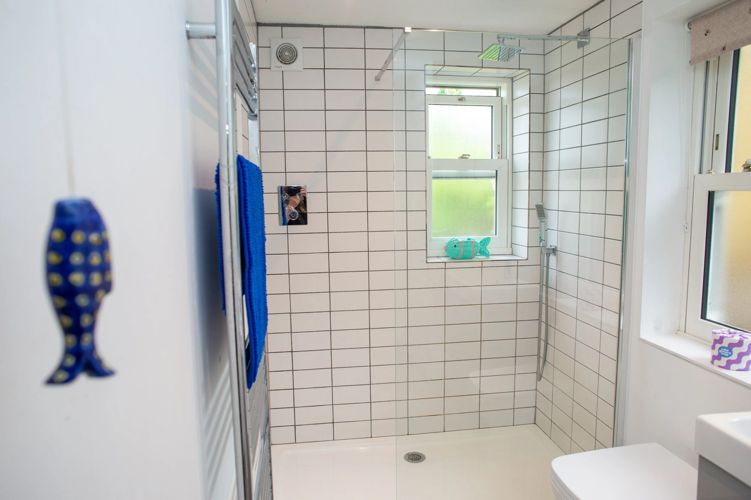 Vintage-style bathroom with a clawfoot bathtub and a wall-mounted sink, both with white porcelain finishes. The bathtub is partially covered with a light blue towel, and a colorful towel hangs from a metal towel rack below the sink. The bathroom has wooden floors and exposed wooden beams on walls and ceiling, with an angled ceiling on the left side.