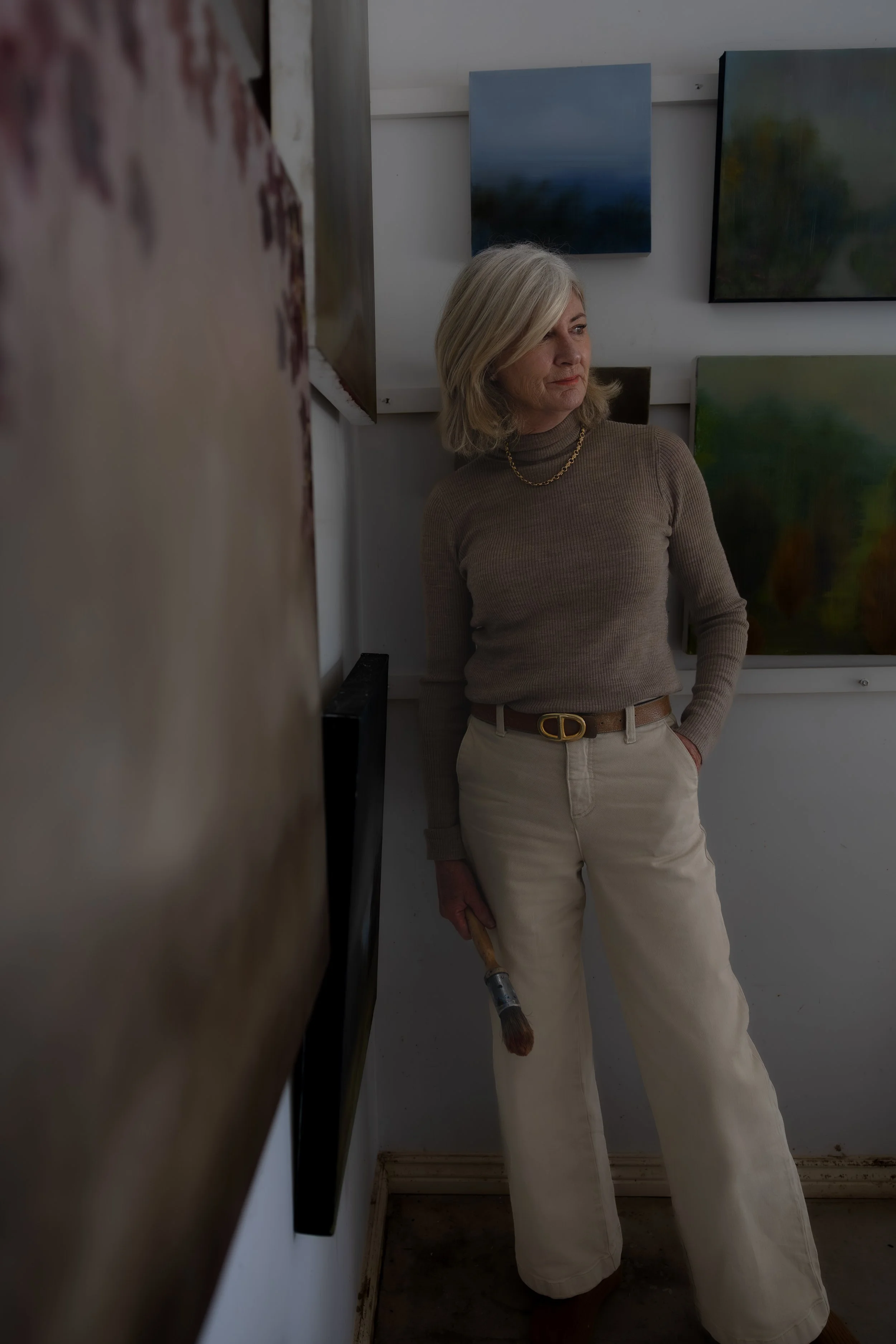 A woman with blonde hair wearing a brown turtleneck and beige pants with a gold belt, standing in an art gallery, holding a paintbrush, surrounded by abstract paintings on the wall.