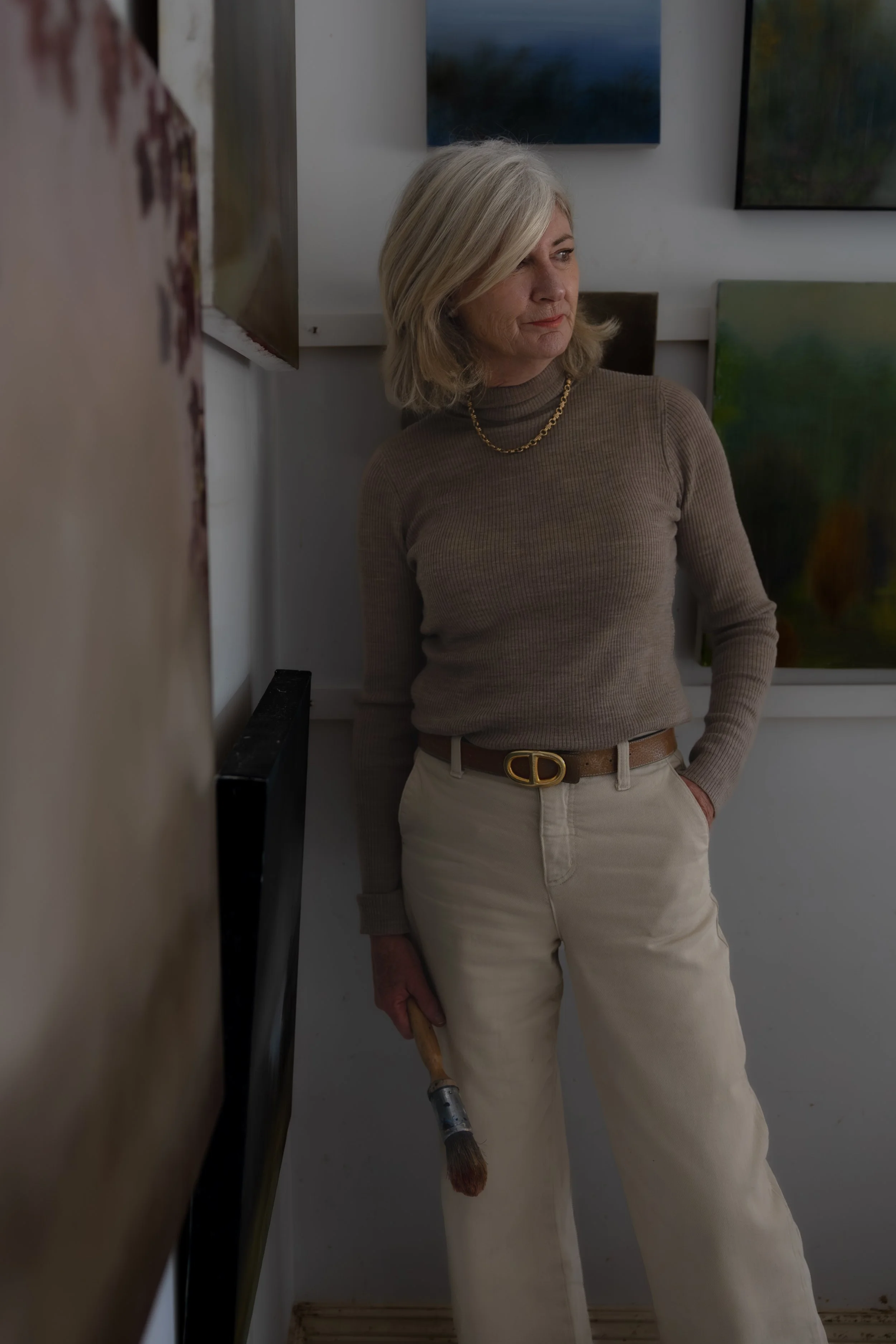 A woman with blonde hair wearing a brown turtleneck sweater, beige pants with a brown belt, standing in an art gallery. She is holding a paintbrush in her right hand and looking to her left. Several paintings are visible on the wall behind her.