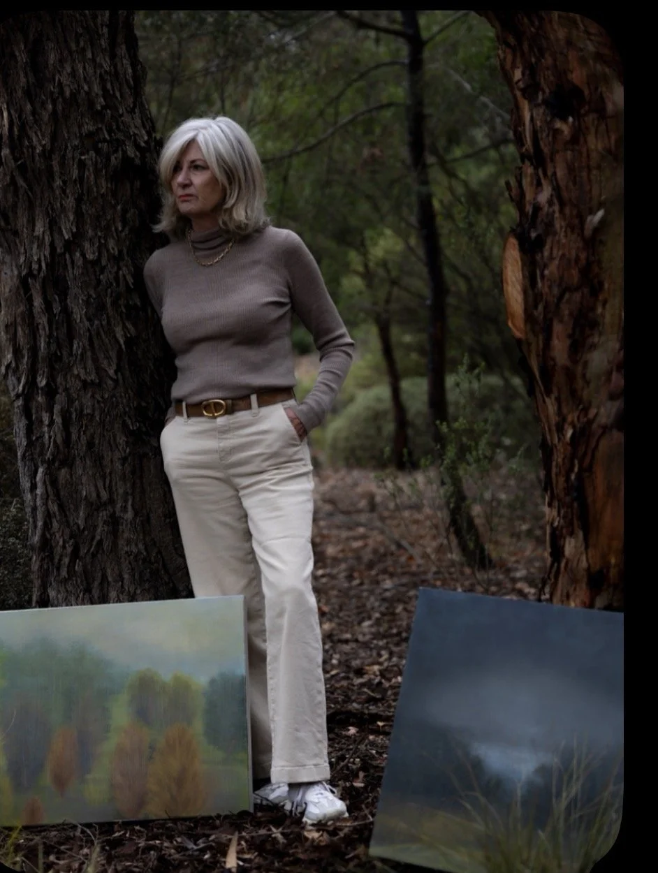 A woman with blonde hair standing in a wooded area, leaning against a tree, with two landscape paintings on the ground in front of her.