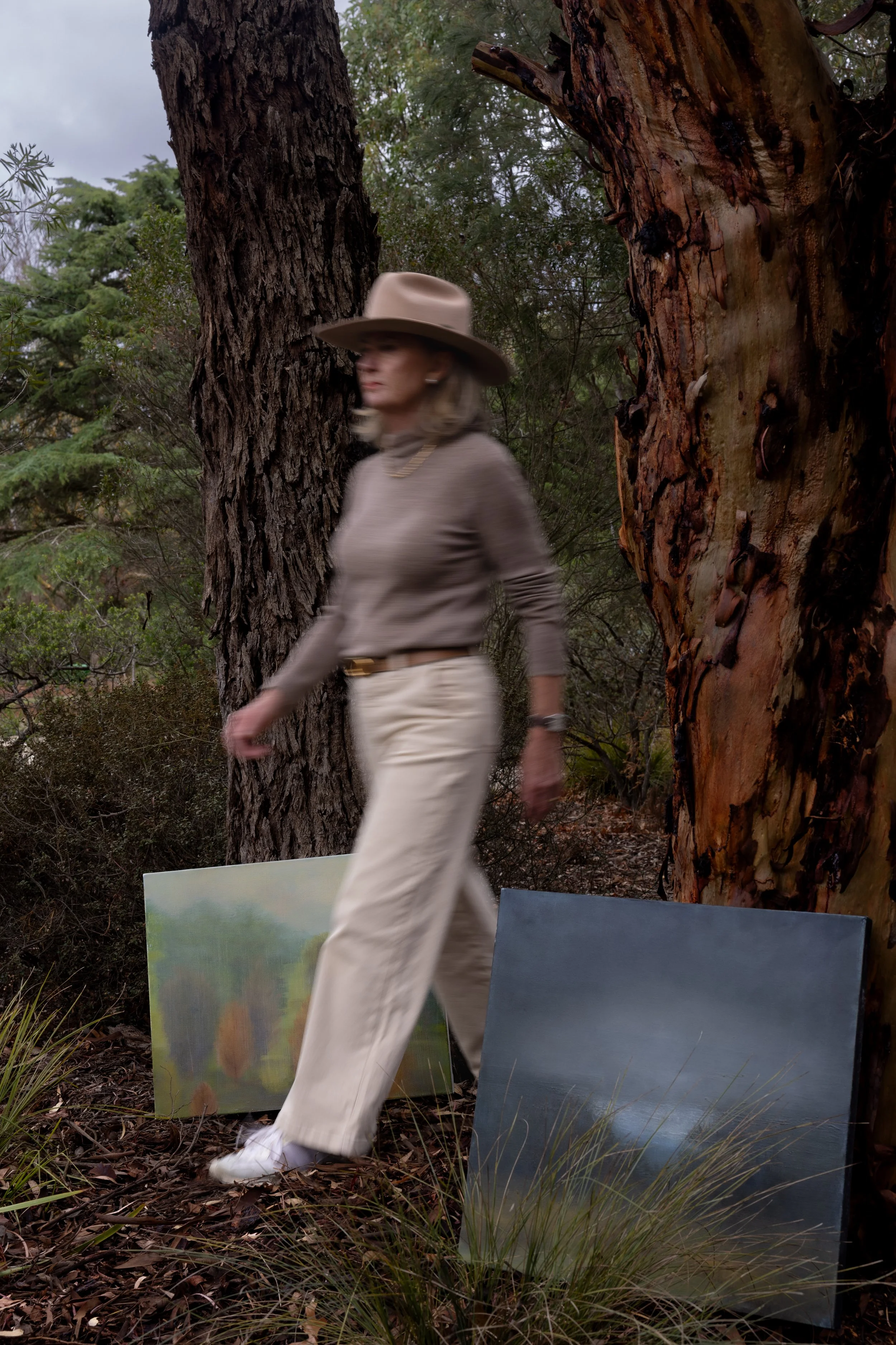 Woman walking through forest area with trees, shrubs, and three landscape paintings on the ground.