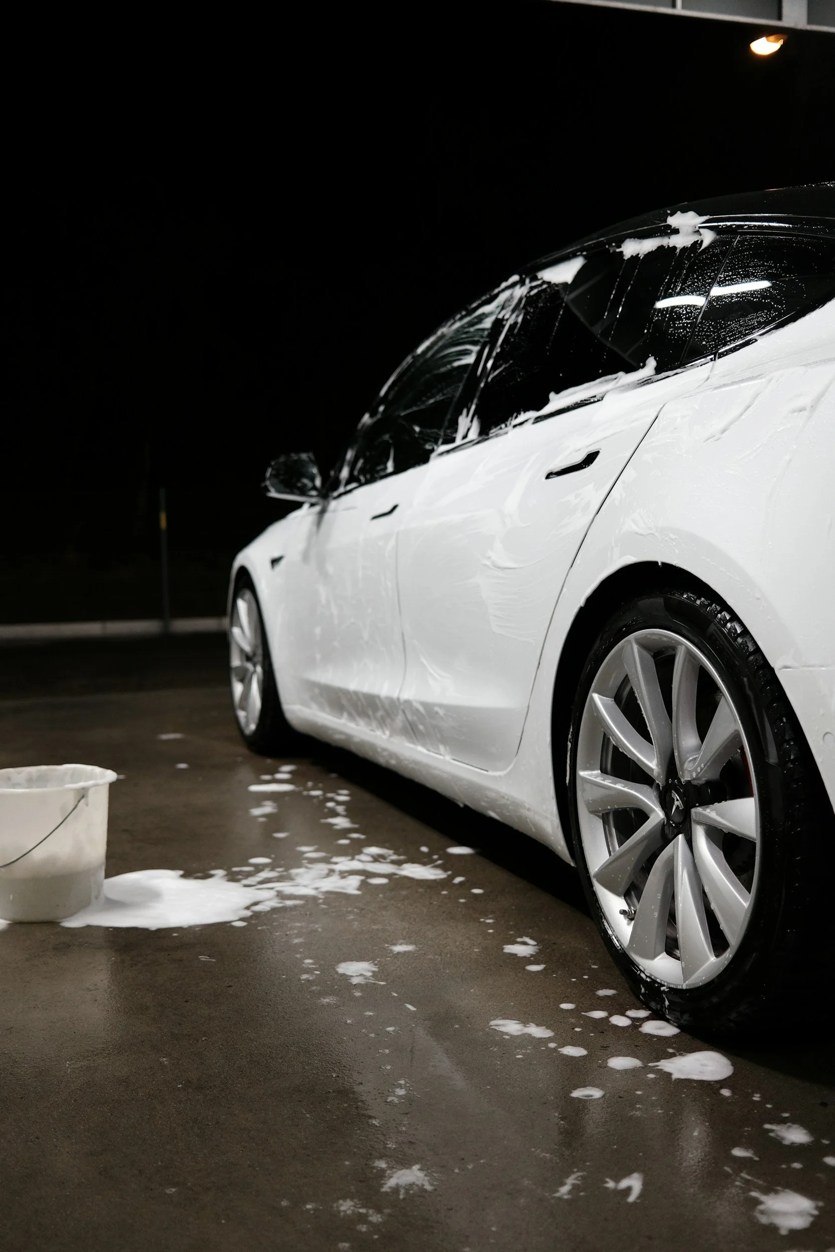 White electric car being washed with soap, foam, and water on a dark night, with a bucket nearby.
