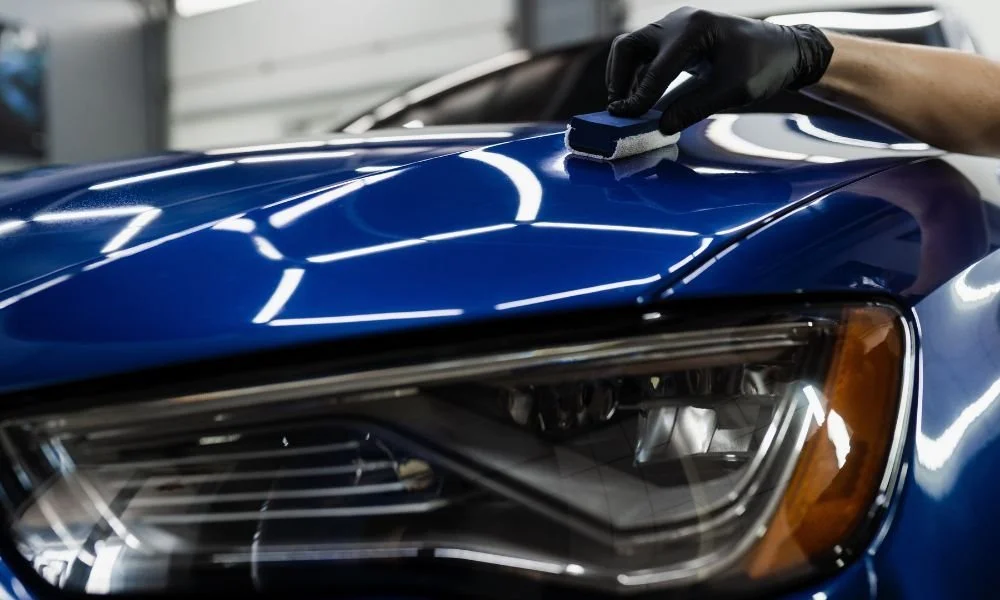 A person wearing black gloves polishing a blue car with a sponge in an indoor garage.