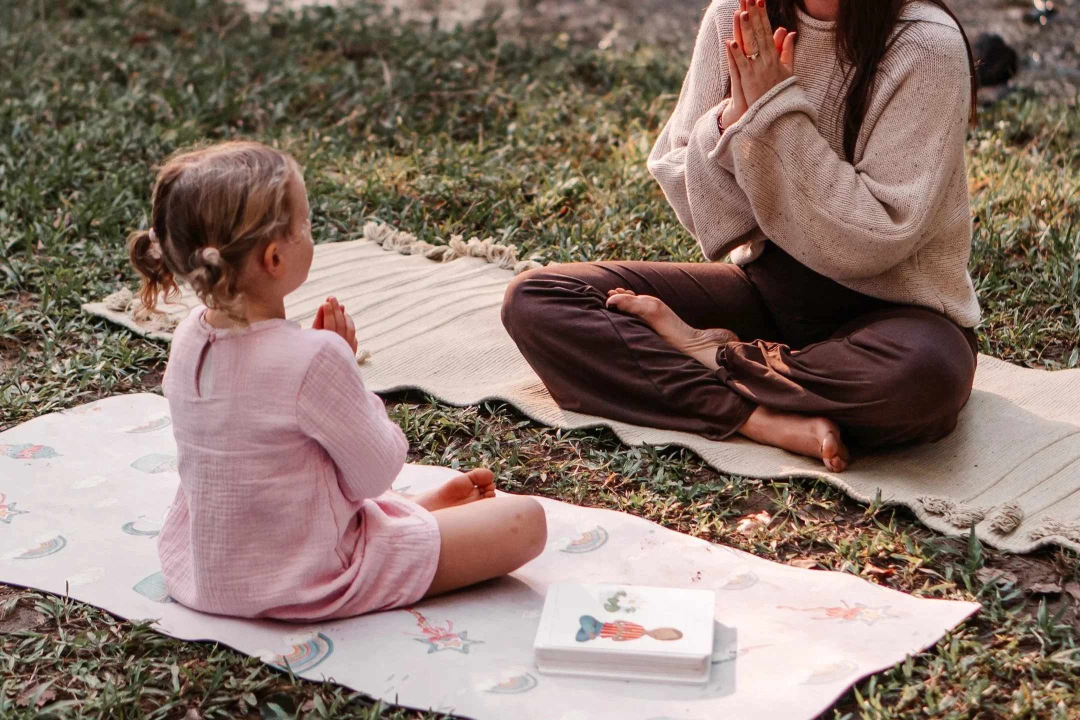 A woman and a young girl sitting outdoors on mats, engaging in a meditation or prayer activity with the girl holding her hands in a prayer position and the woman with her hands in a prayer position, surrounded by grass and fallen leaves.