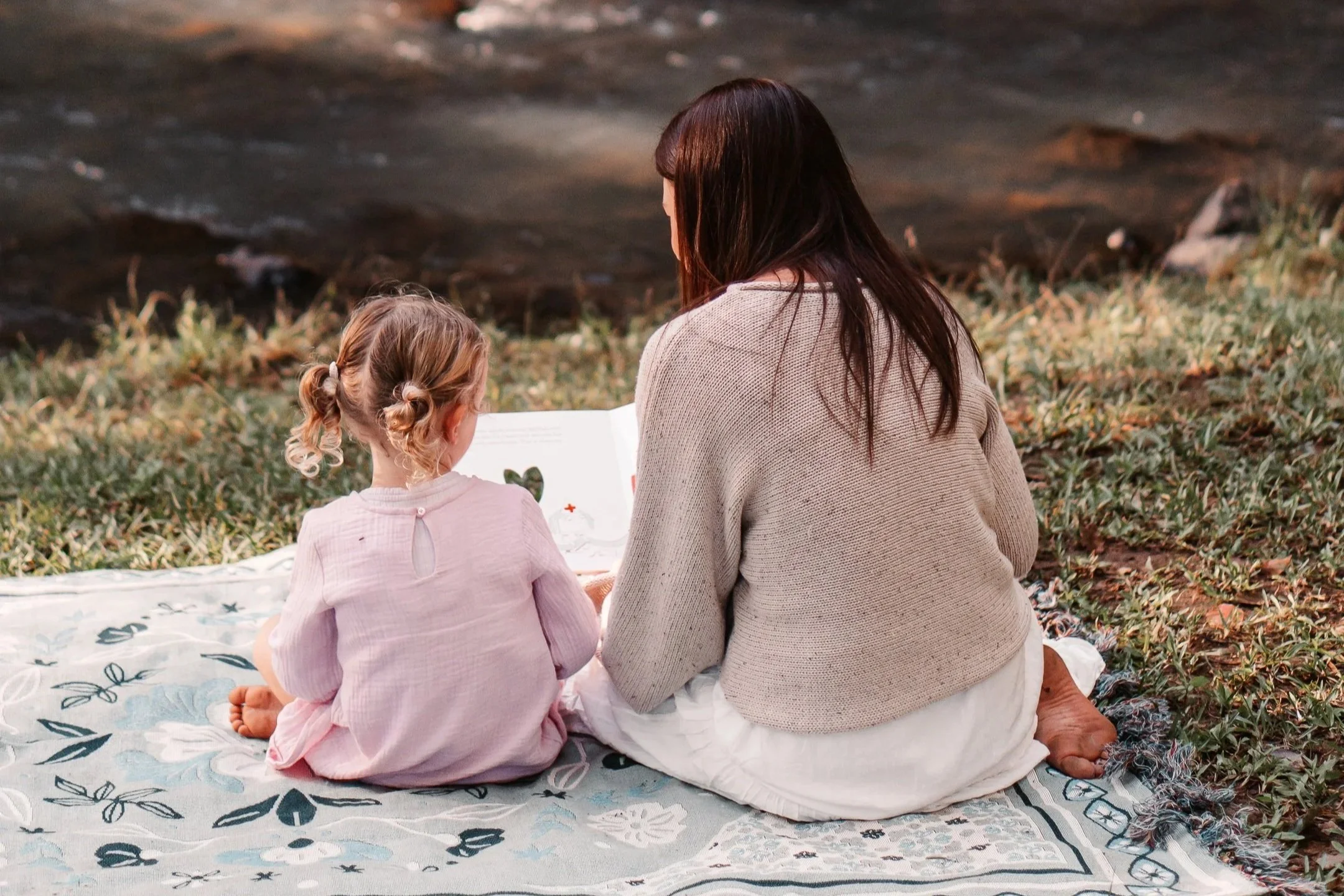 A woman and a young girl sitting on a blanket by a body of water, facing away from the camera.