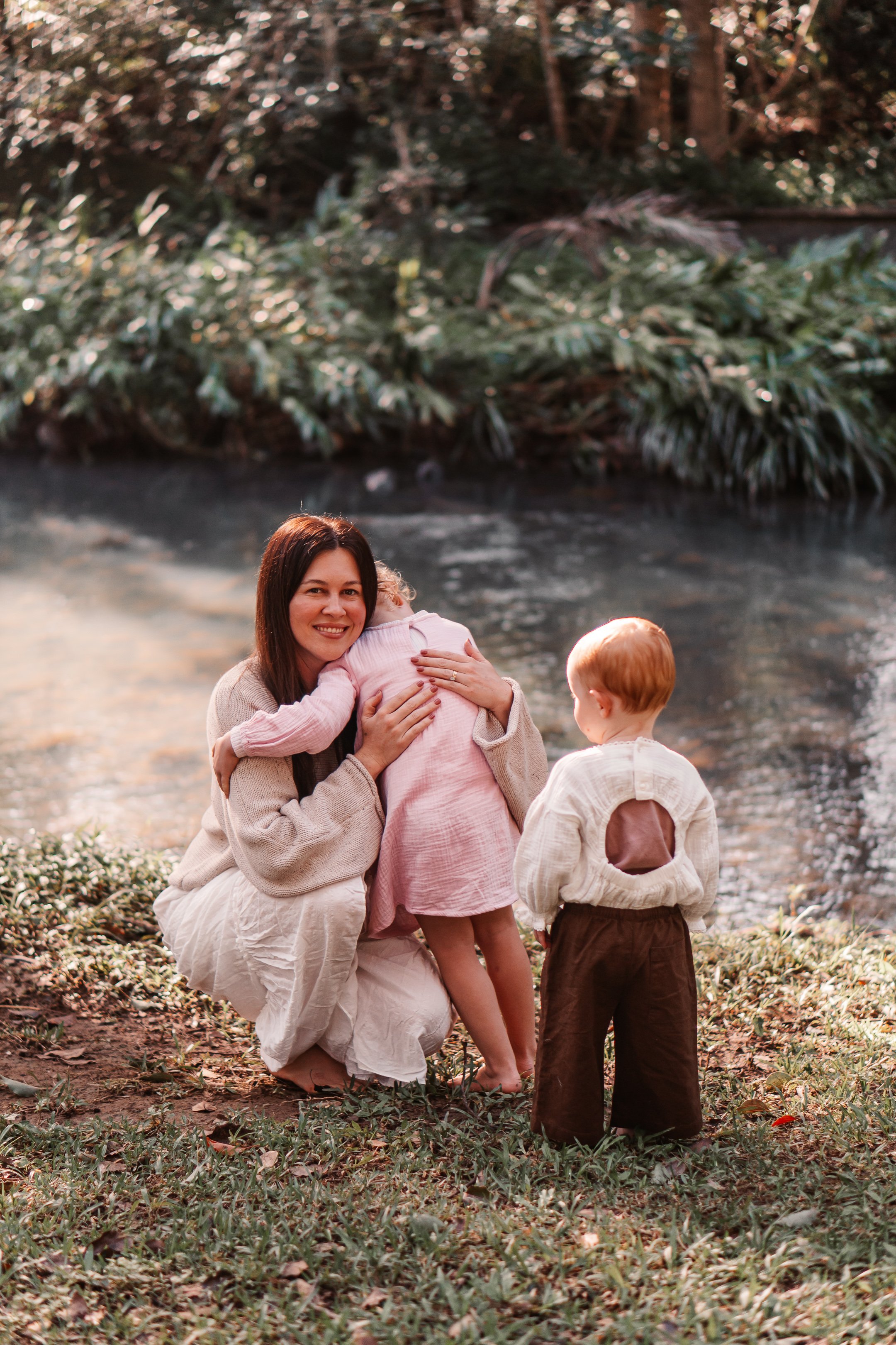 A woman kneeling on the ground by a river, hugging a small girl in a pink dress. A boy with red hair and brown pants stands nearby, facing the river. The background features trees and foliage, suggesting an outdoor, natural setting during daylight.