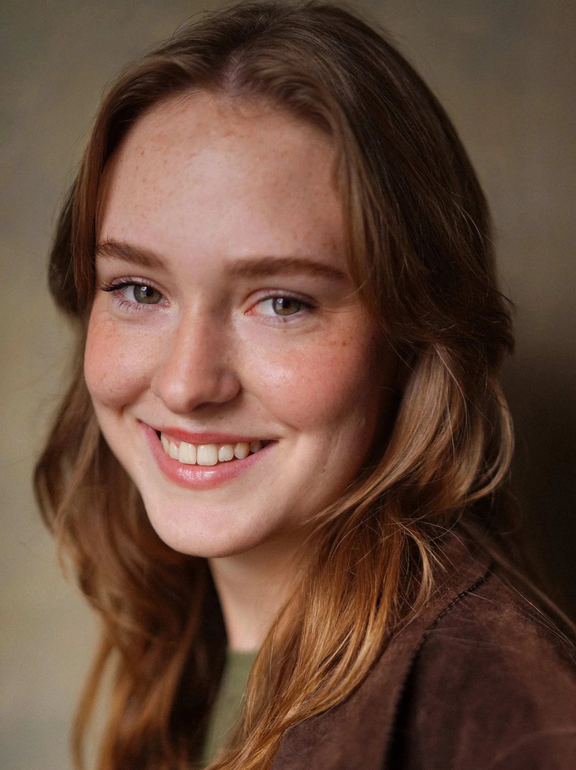 Close-up of a young woman with long, wavy auburn hair, freckles, light eye makeup, and a friendly smile, wearing a brown leather jacket.