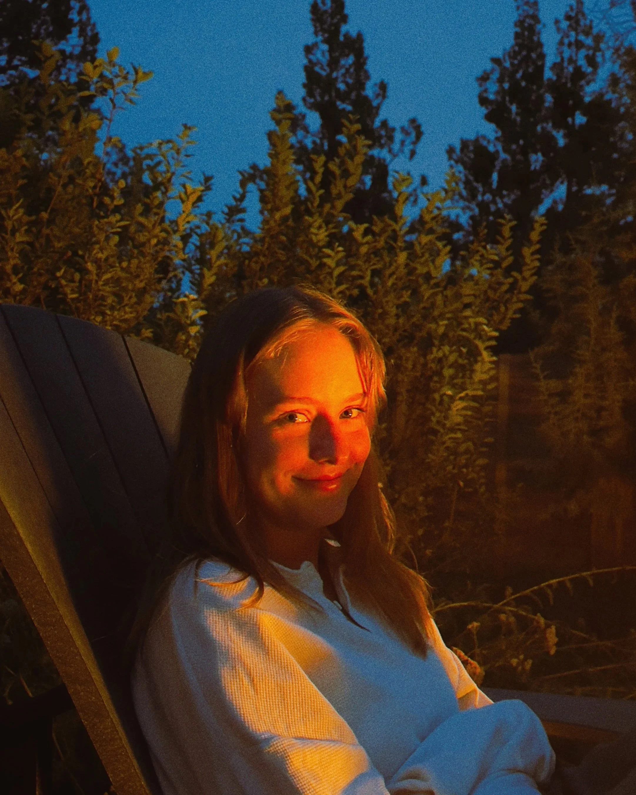 A smiling person with long hair sitting outdoors at dusk, backlit by warm light, with trees and the evening sky in the background.
