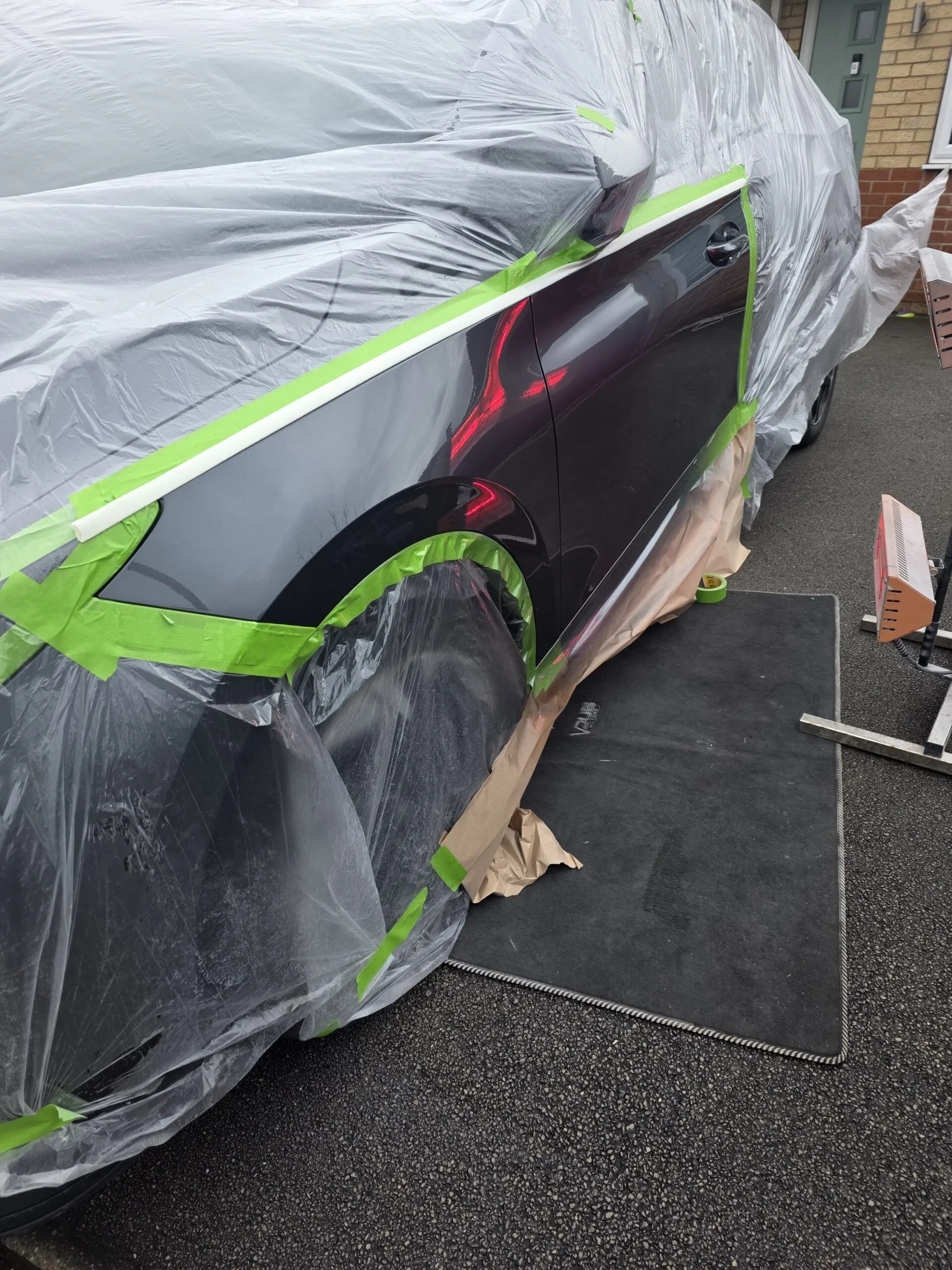 Une voiture en cours de peinture, protégée par un film plastique, avec des bandes de masquage vert et du papier brun pour protéger certaines parties