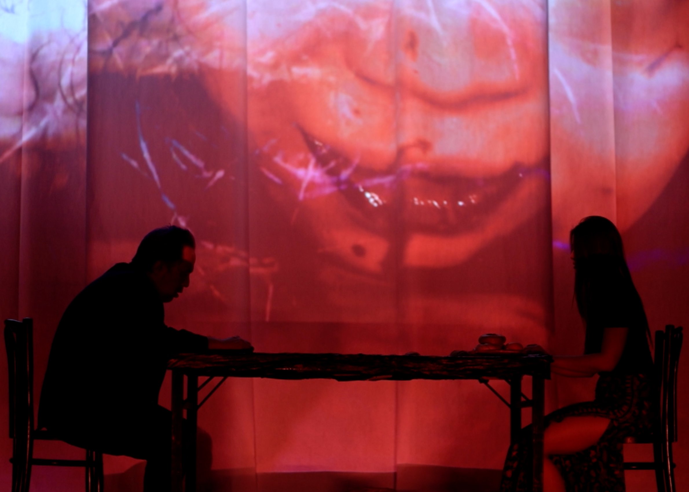 Performance still from Prayers for a Hungry Ghost (2025).  Two silhouetted figures sit at opposite ends of a table  in deep red light, a large projected close-up of a face  looming behind them on a screen