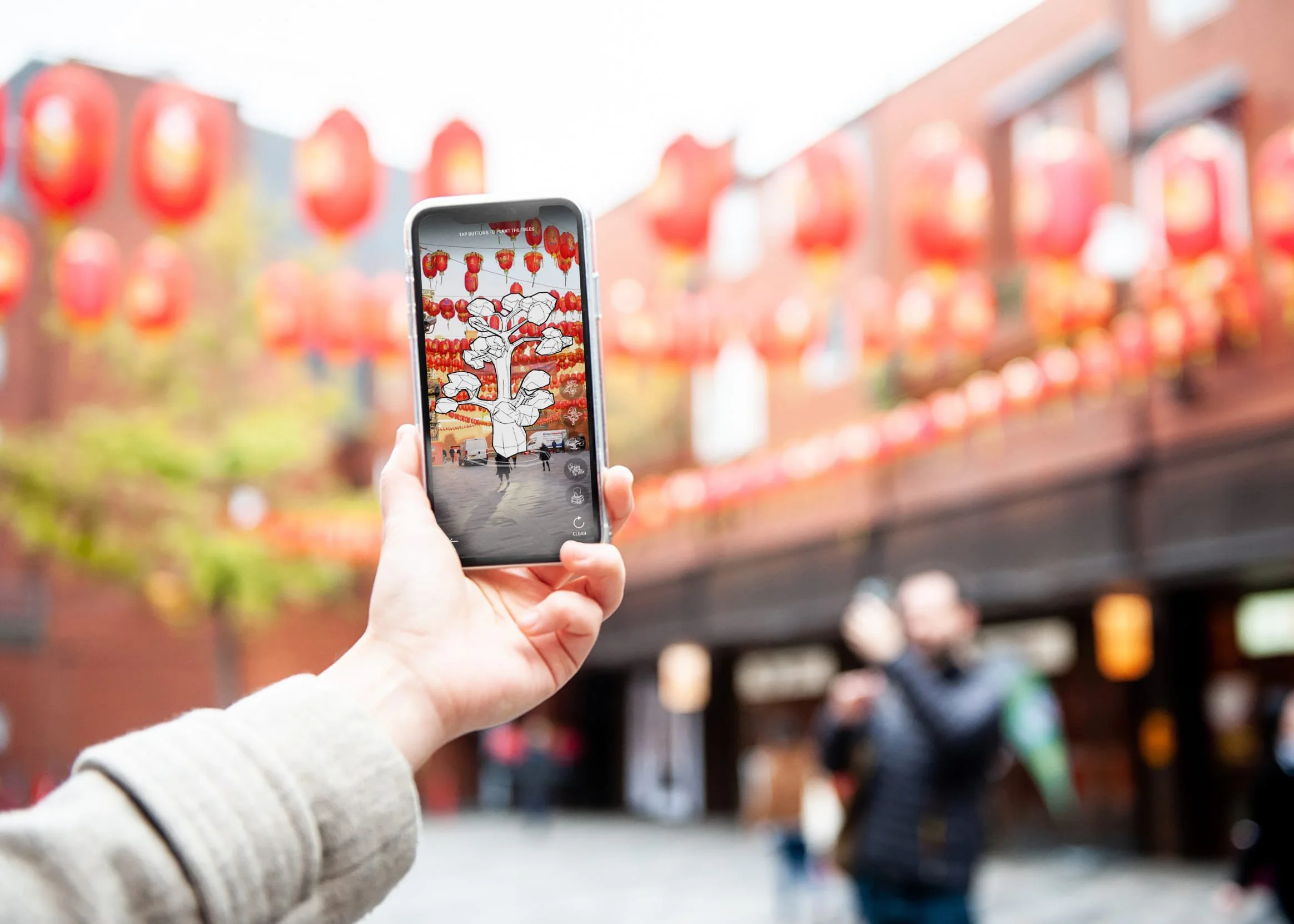 Documentation of Augmented Chinatown (2022). A hand holds  a smartphone displaying an AR tree overlaid onto London's  Chinatown, with red lanterns strung above the street  visible in the background