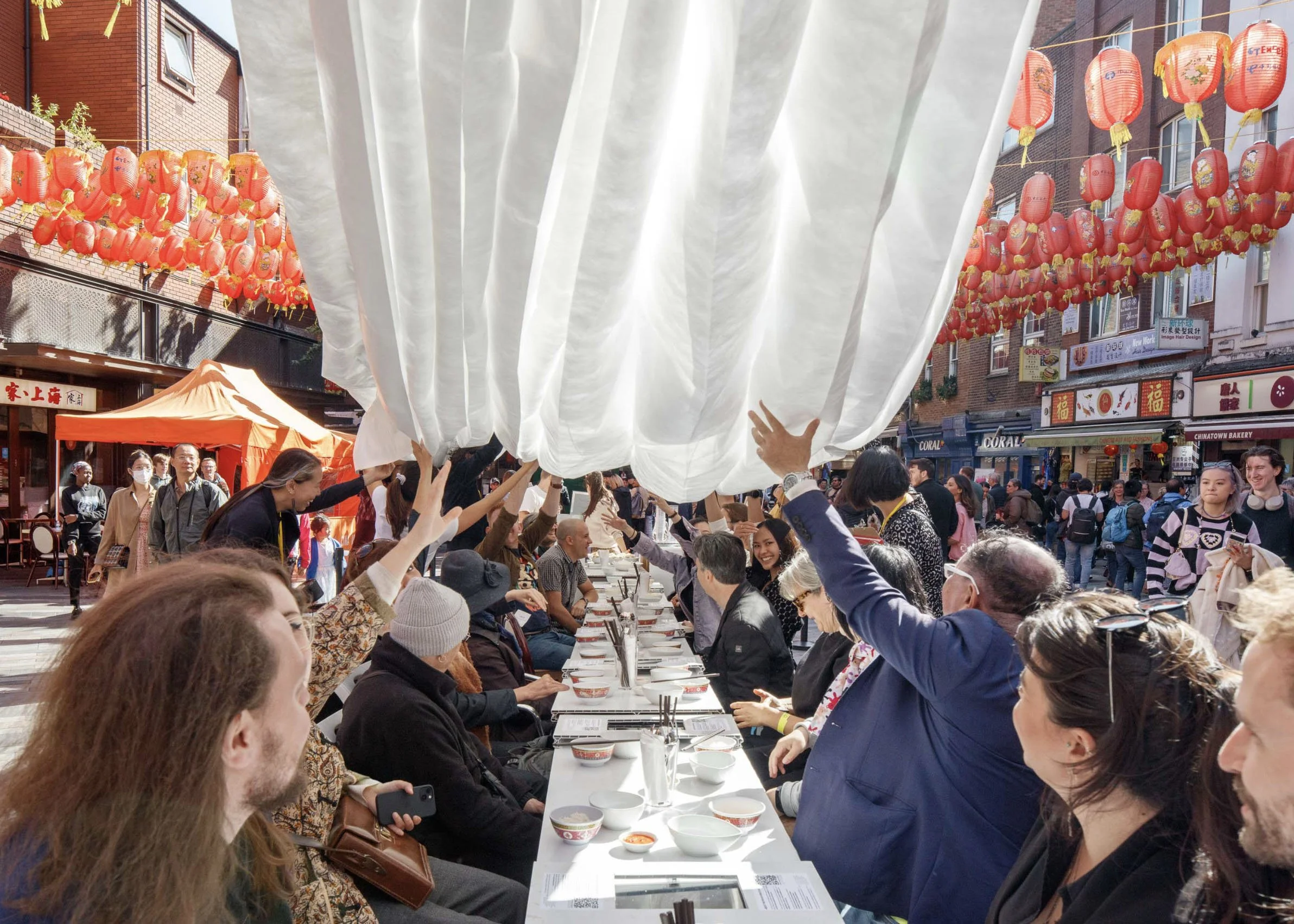 Documentation of Low Carbon Chinatown. A large crowd  seated at a long communal table on a London Chinatown  street raise their hands to billow a white fabric sheet  overhead, surrounded by red lanterns, shopfronts, and  onlookers on a sunny day