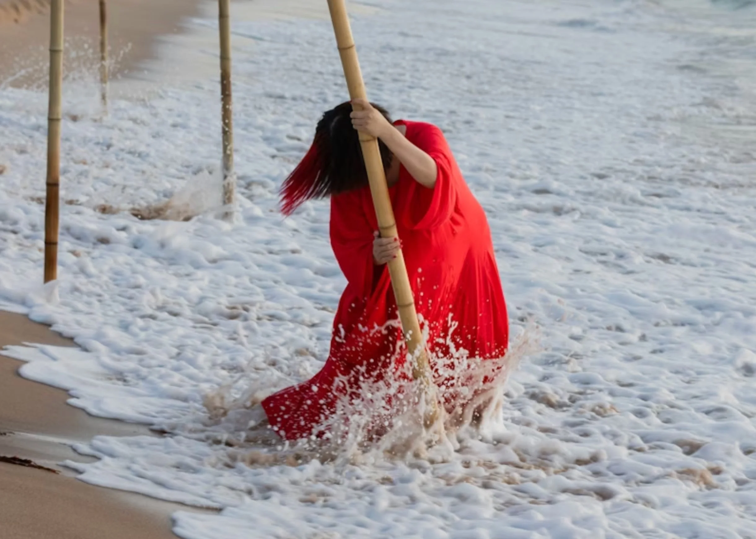 Performance still from Xiao Lu. A performer in a  sweeping red dress grips a bamboo pole amid crashing  ocean waves on a beach, head bowed against the surge,  surrounded by other bamboo poles standing in the surf.