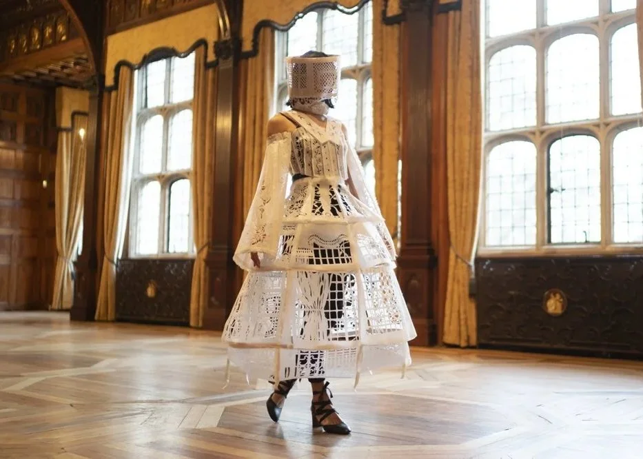 Documentation of artwork by Chloe Wing at Two Temple  Place (2021). A figure wearing Chloe Wing's elaborate  white laser-cut costume and face-obscuring headpiece  stands in a grand wood-panelled hall with tall arched  windows and parquet flooring