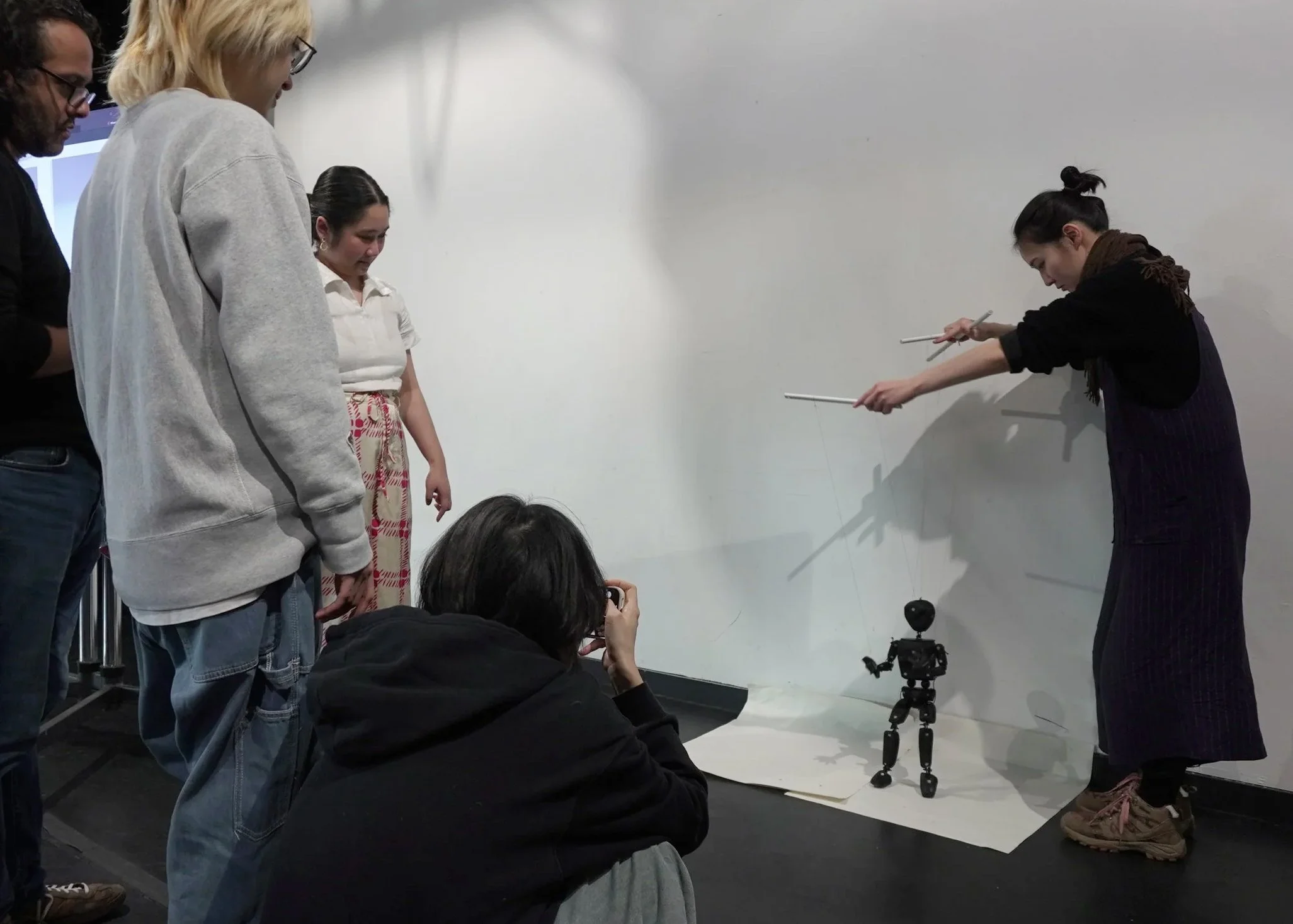 A puppeteer manipulates a small black articulated robot puppet with  control rods while three audience members watch closely,  one crouching to photograph, casting dramatic shadows  on a white gallery wall