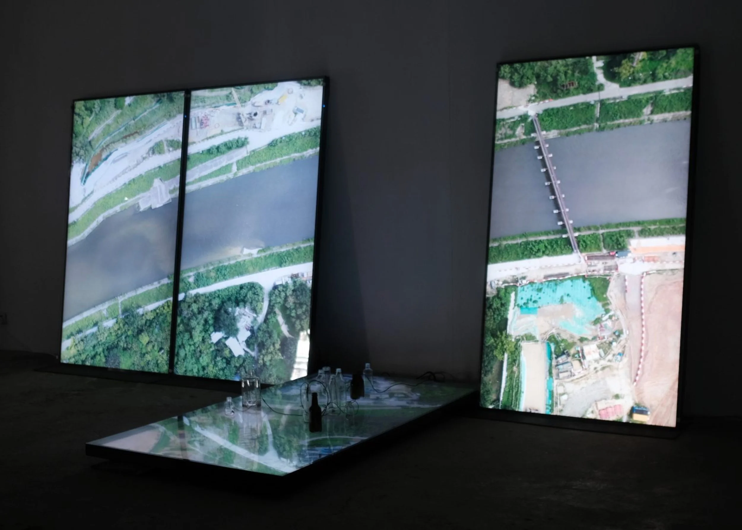 Installation view of State/Less (2023). Three large vertical  screens display aerial footage of rivers and urban terrain  in a darkened gallery, with a low illuminated table holding  bottles and objects in the foreground