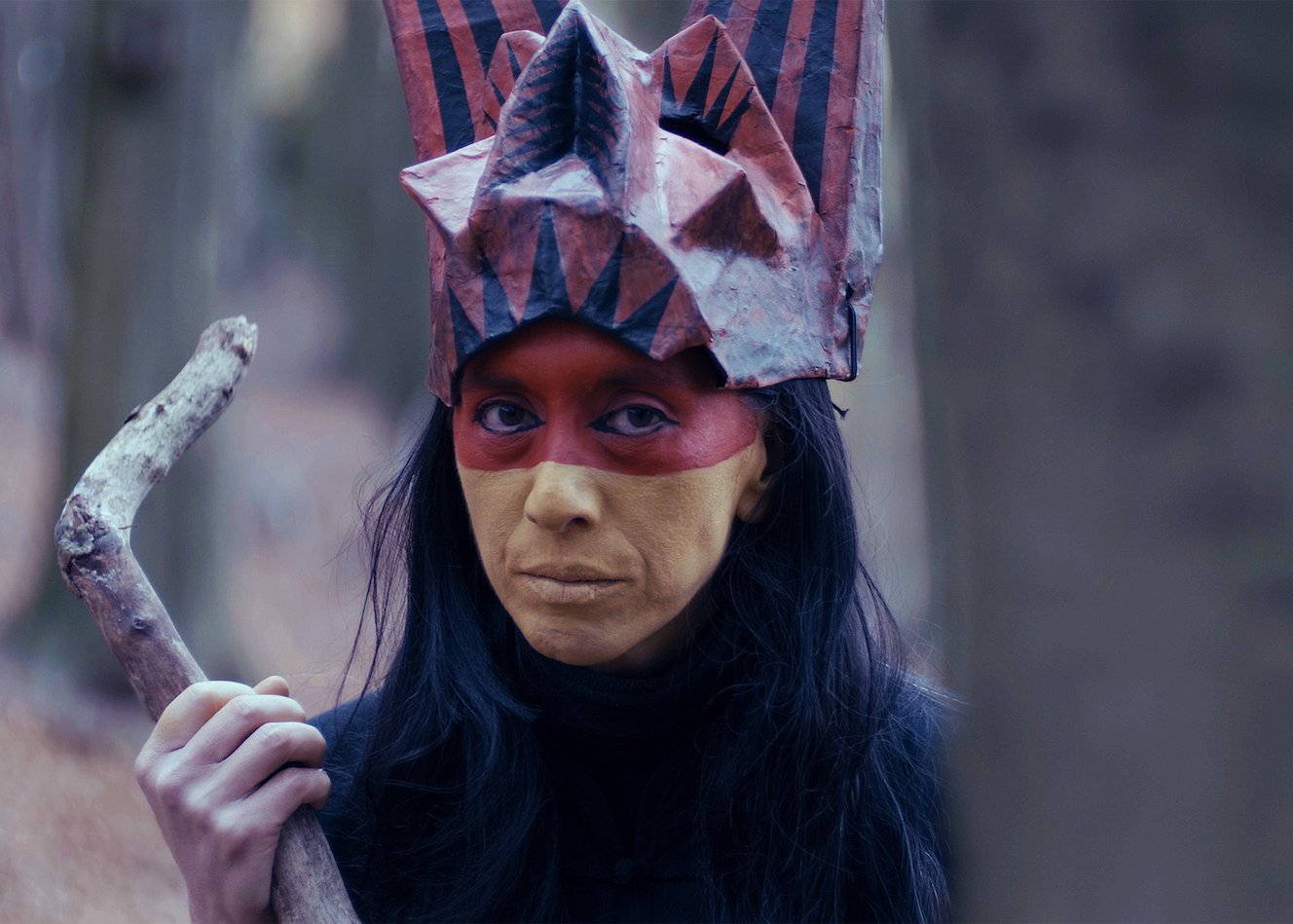 Film or performance still from Casting Fu Manchu (2021).  A theatre artist wearing red and gold face paint and a  handmade geometric paper crown holds a gnarled branch,  gazing directly at the camera in a woodland setting