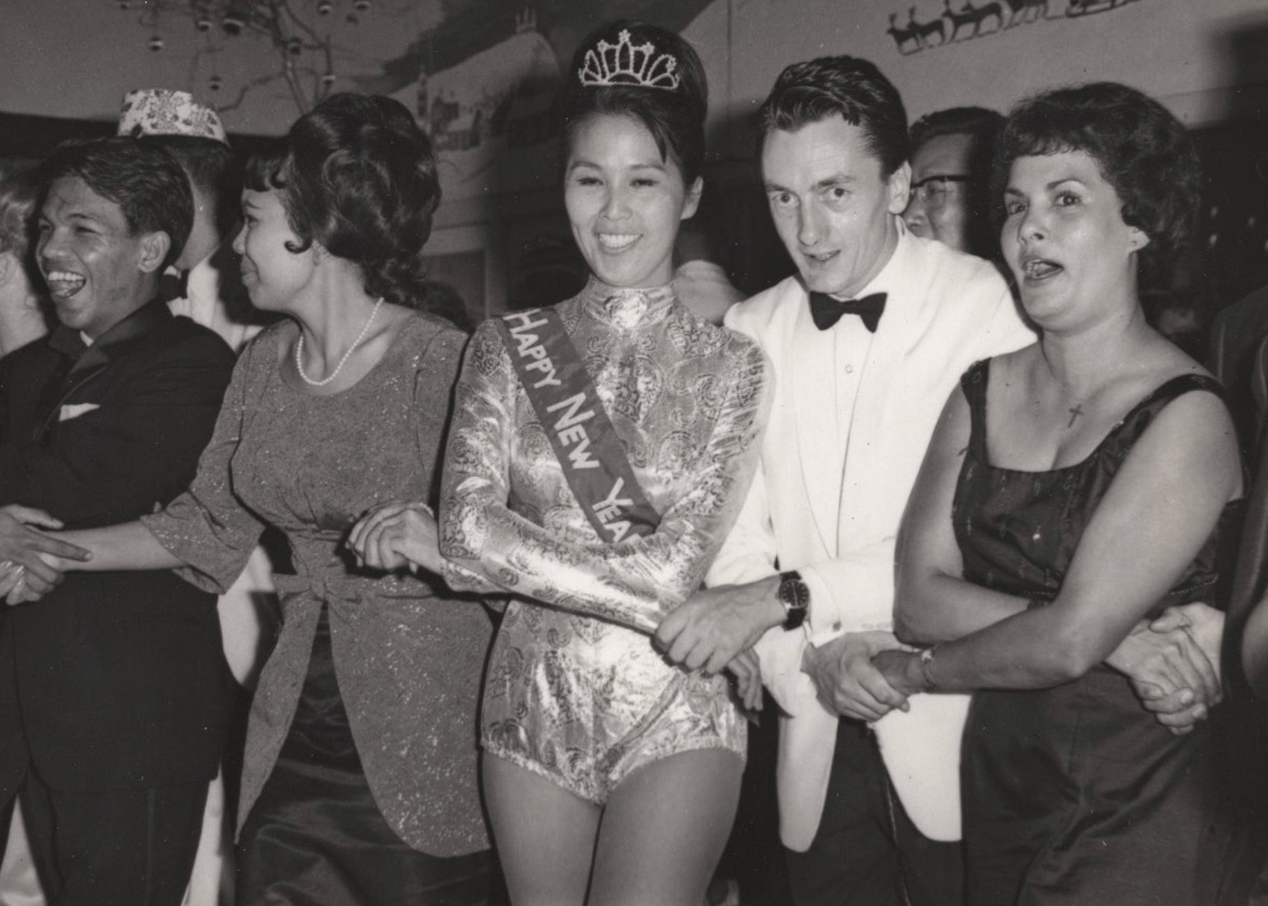 Archival photograph used in Duck Rabbit. A woman wearing  a tiara, brocade bodysuit and "Happy New Year" sash  dances in a conga line with a mixed crowd at what appears  to be a New Year's celebration, photographed in black  and white, circa 1960s