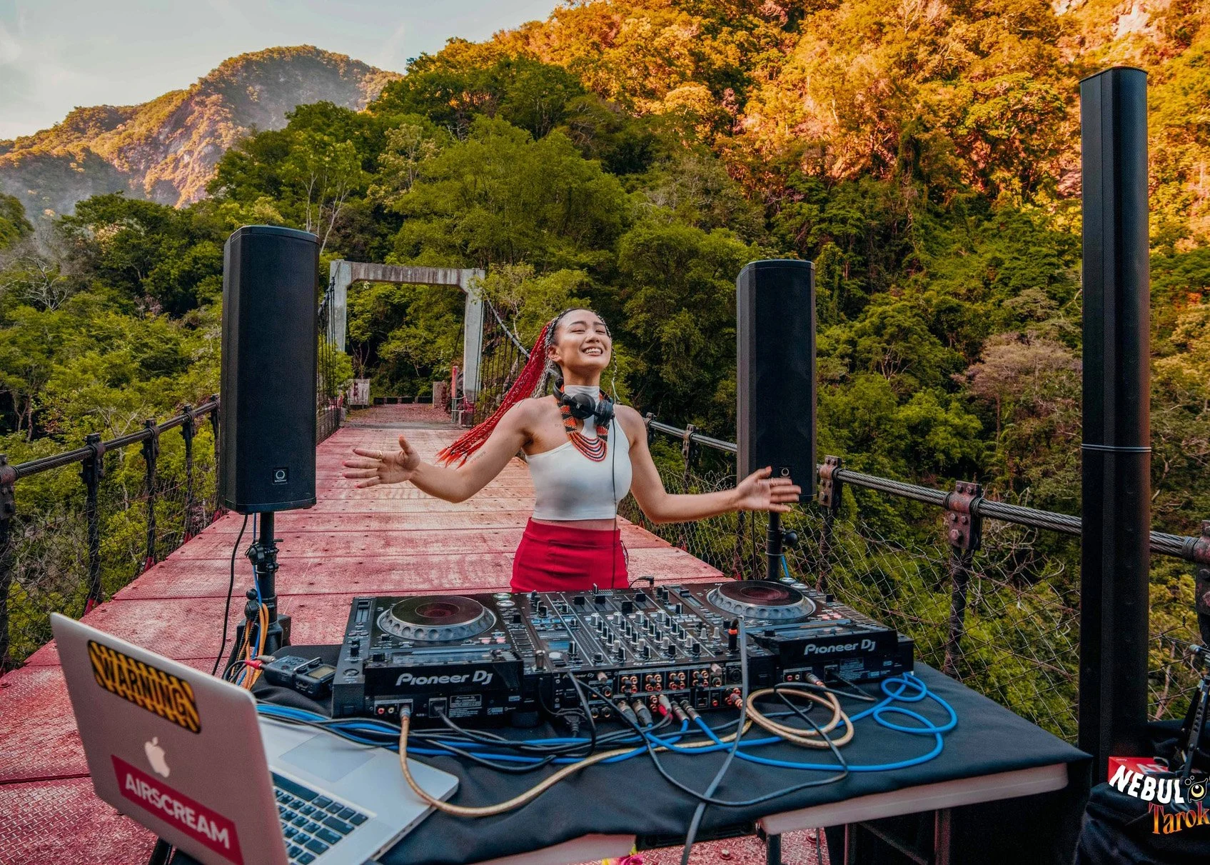 DJ Dungi performs at DJ decks on a suspension bridge  surrounded by lush forested mountains, wearing traditional  Taiwanese Amis costume including red braided hair  extensions and beaded necklace, arms outstretched  and smiling at golden hour