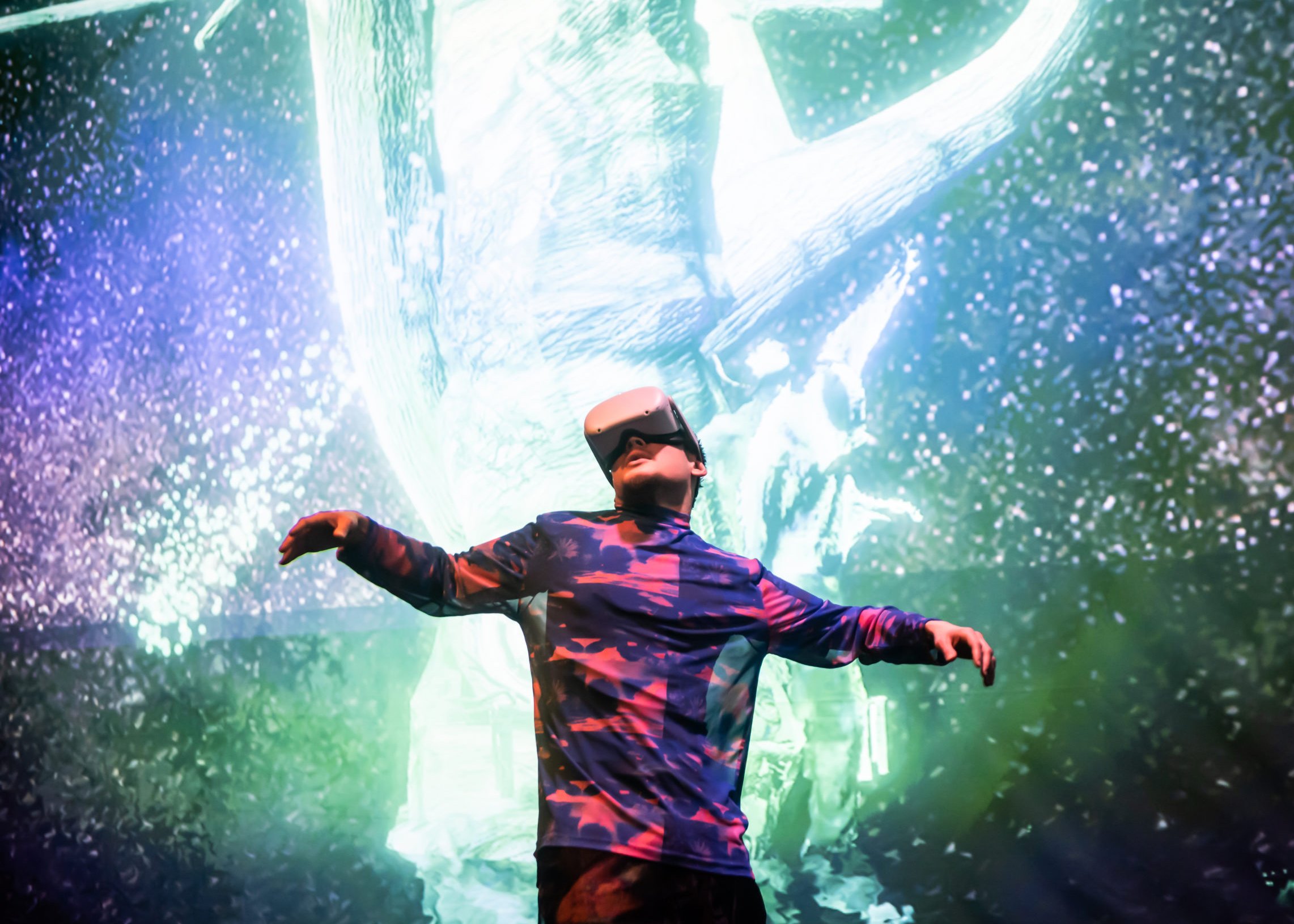 Performance or installation documentation from Home X.  A figure wearing a VR headset stands with arms outstretched  amid an immersive floor-to-ceiling projection of a luminous  forest environment in teal and purple
