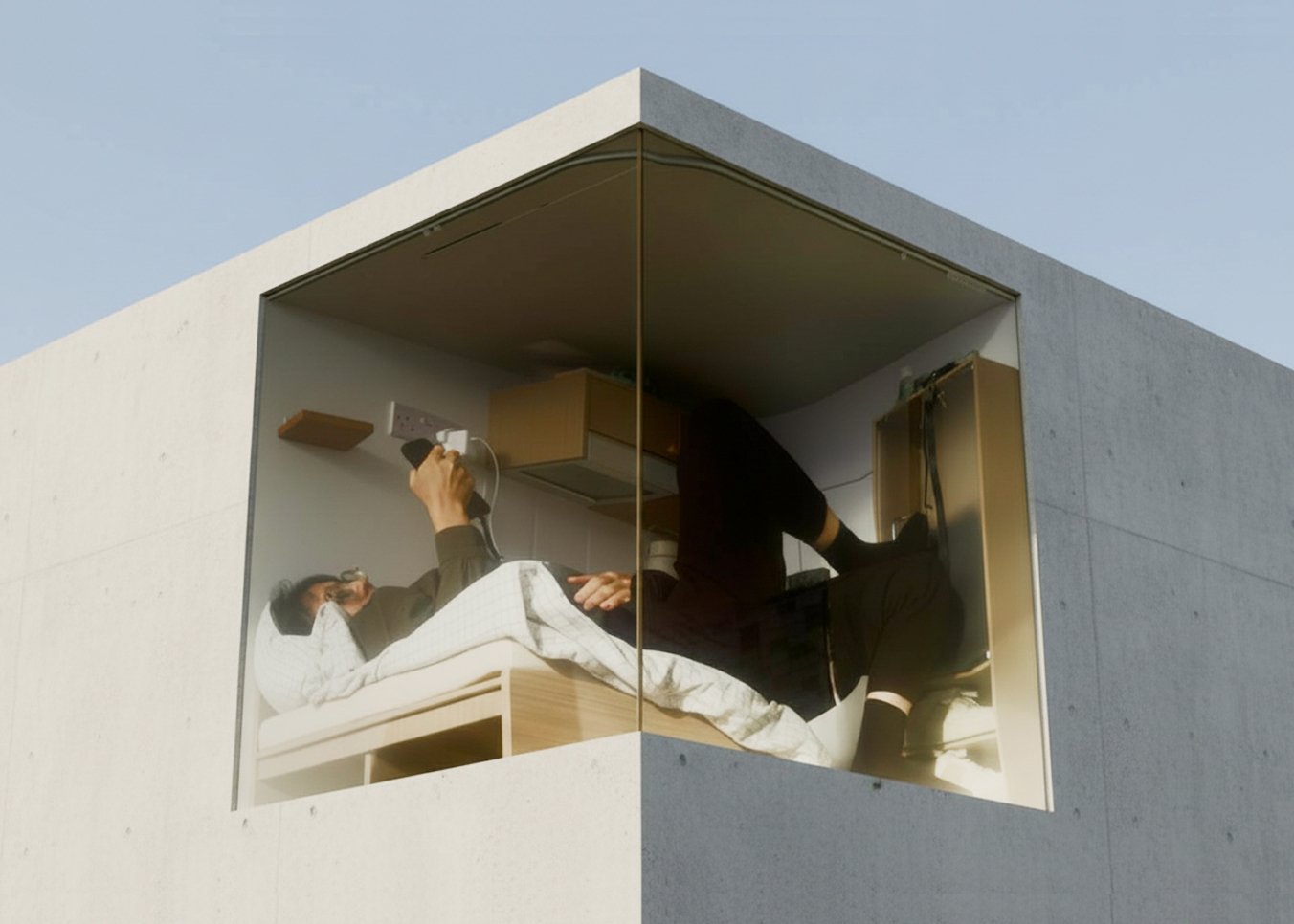 Concept image for PCM (2025). A figure occupies a cramped  concrete pod elevated against a pale sky — lying on a  narrow bed holding a hairdryer, legs extending through  a doorway, viewed through a corner window cut into  the structure