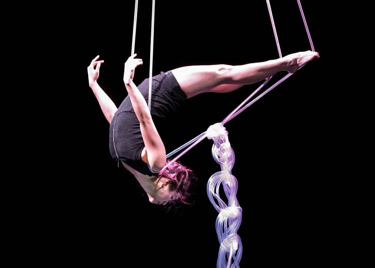 Performance still from Light Vessel. An aerial artist  suspended upside down in straps, body arched and legs  extended, holding a luminous braided rope of light  tubes against a black background