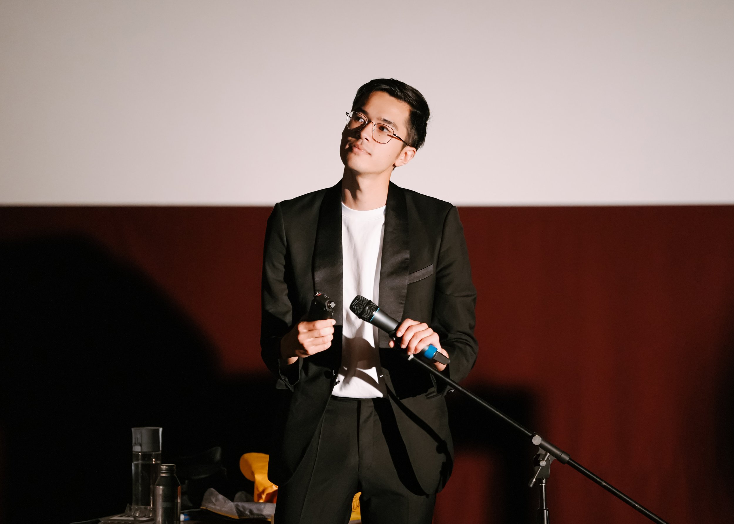 Theatre artist holding a microphone on stage before a cinema screen and deep red wall, gazing upward in a moment of reflection
