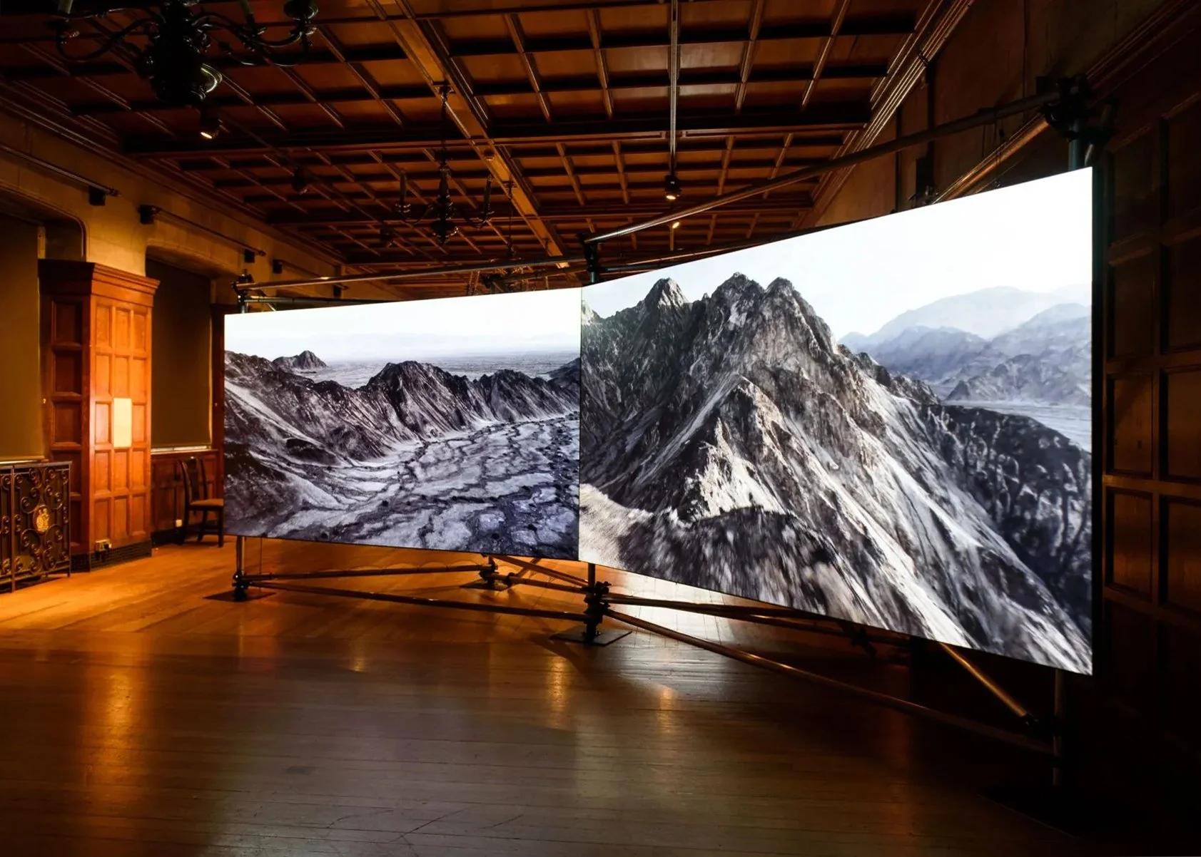 Installation view of State/Less (2023). Two large curved  screens display sweeping aerial footage of arid mountain  ranges in monochrome, installed in a grand wood-panelled  hall with ornate ironwork and a coffered ceiling