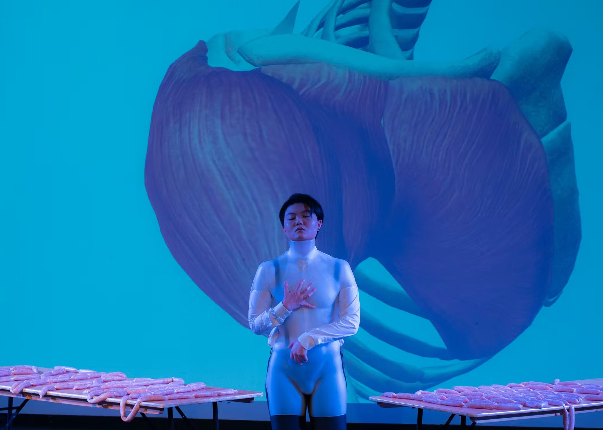 Performance still from Queering Now (2021). A theatre  artist in a white bodysuit stands with eyes closed and  hand on chest before a large projected close-up of a  human heart in teal light, flanked by tables lined  with sausages