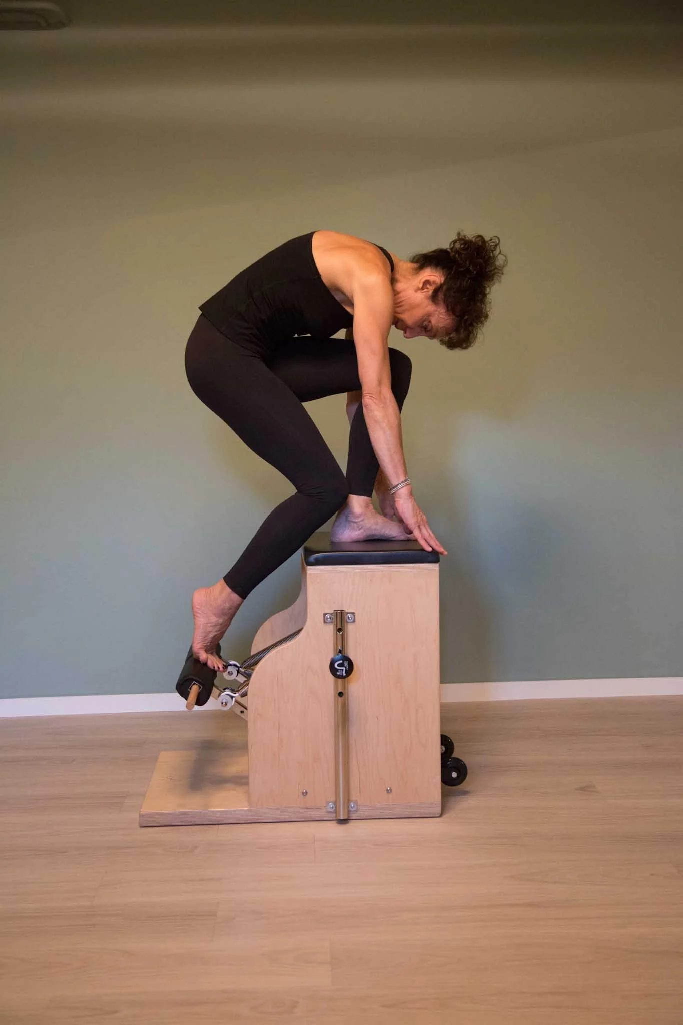 Woman in black workout clothes performing a Pilates exercise on a wooden Pilates chair.