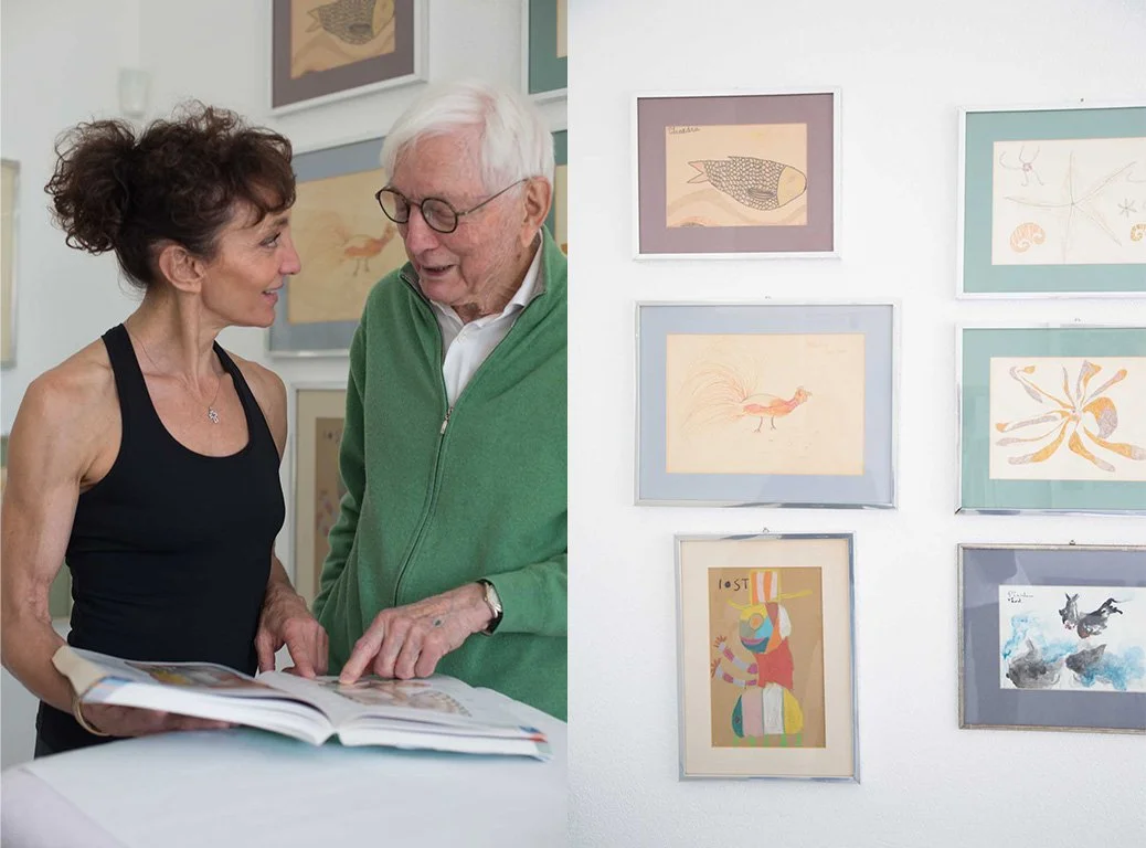 Older man and woman reviewing an art book beside a wall displaying framed artwork.