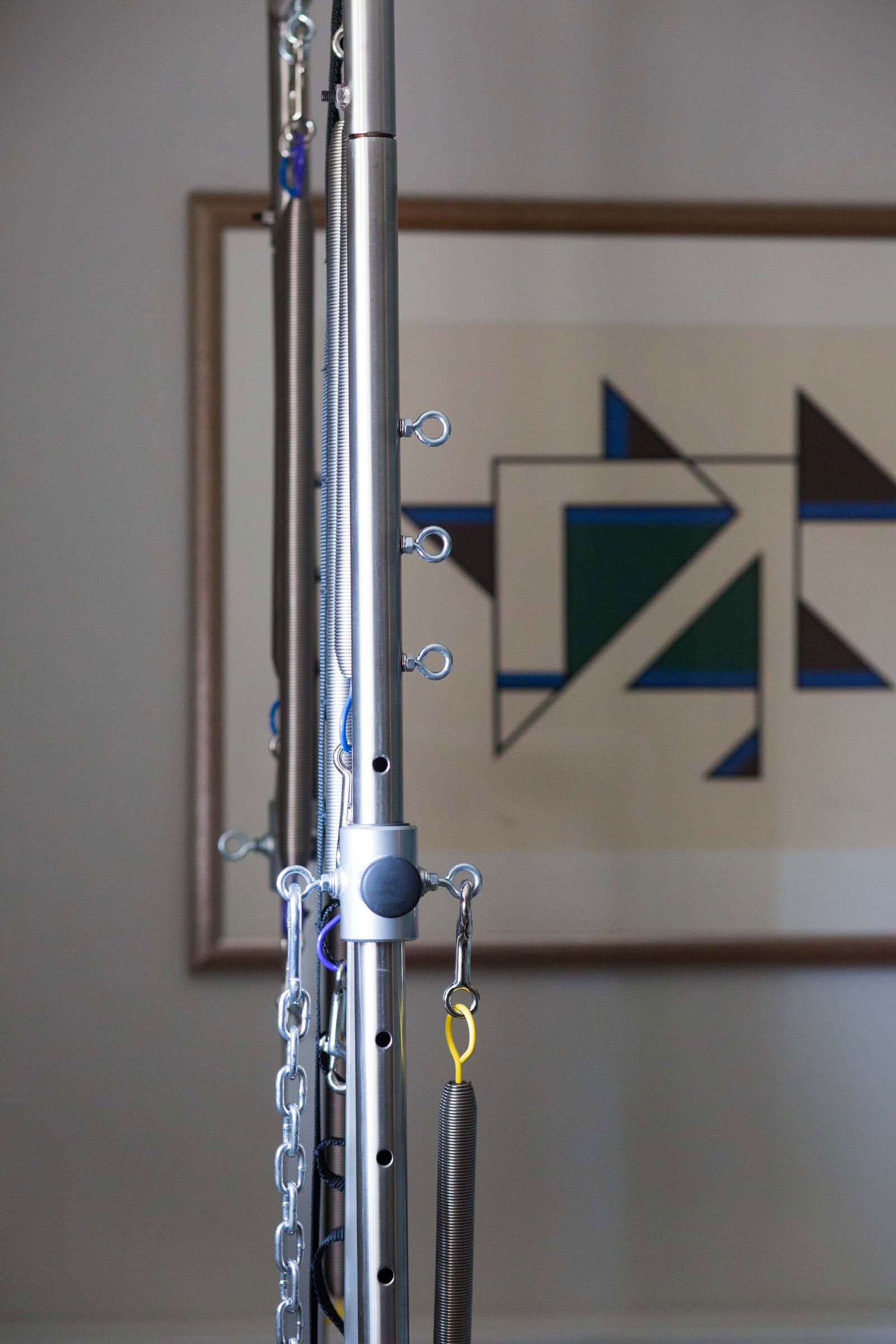 Close-up of a silver aerial dance or fitness rig with springs, chains, hooks, and adjustable holes in front of geometric wall art