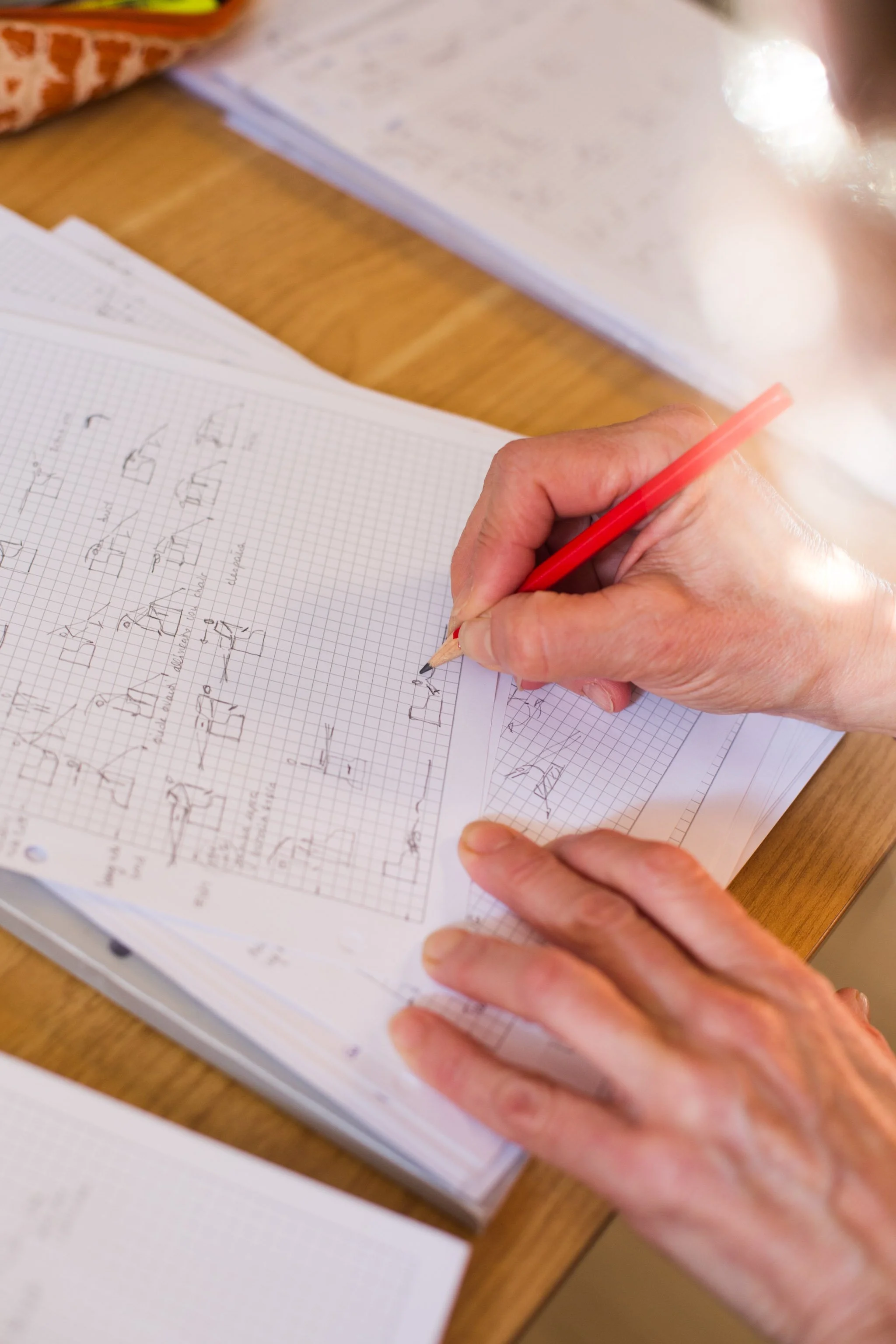 Hands sketching architectural designs in a graph-paper notebook with a red pencil on a wooden desk