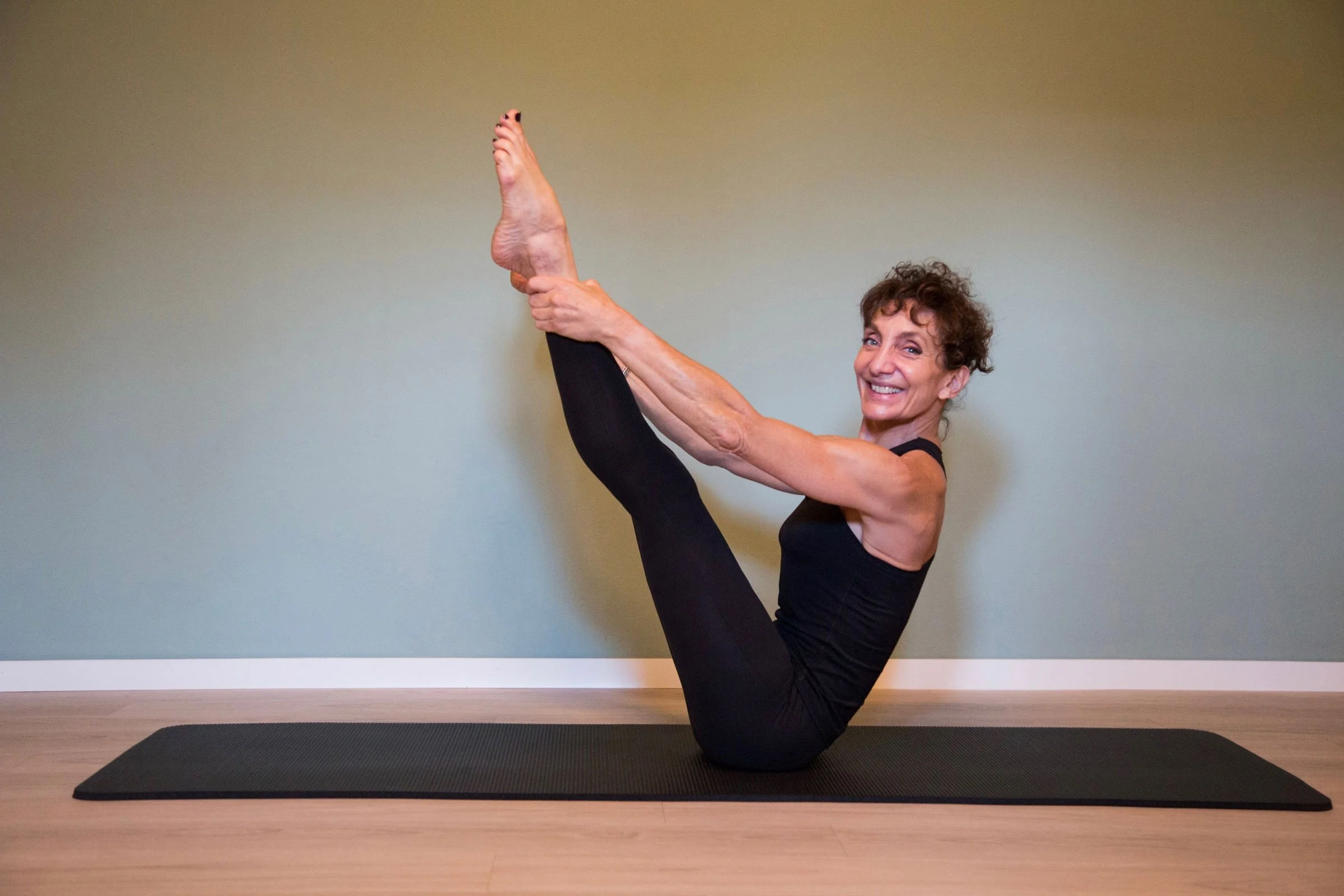 Woman in black workout clothes performing a seated yoga stretch on a mat, holding one raised leg.