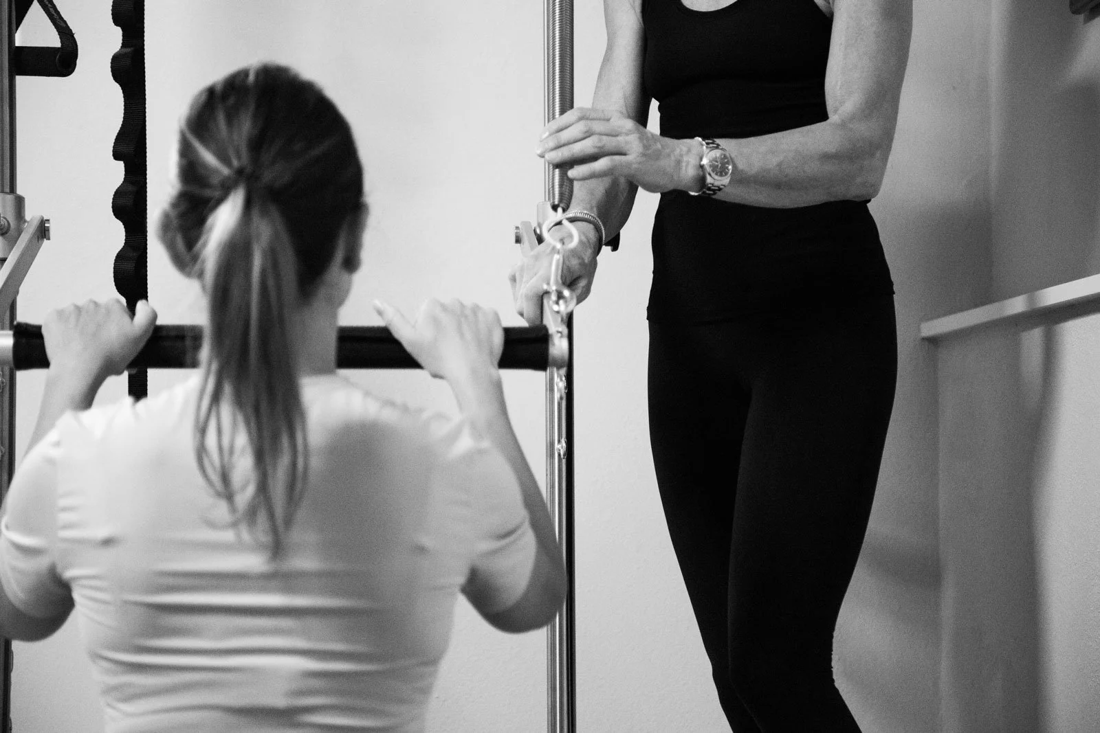 Black-and-white photo of a woman exercising on a Pilates reformer with an instructor assisting her.