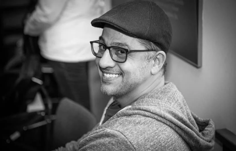 Smiling, friendly man wearing glasses and a flat cap, sitting indoors, with a blurred background.
